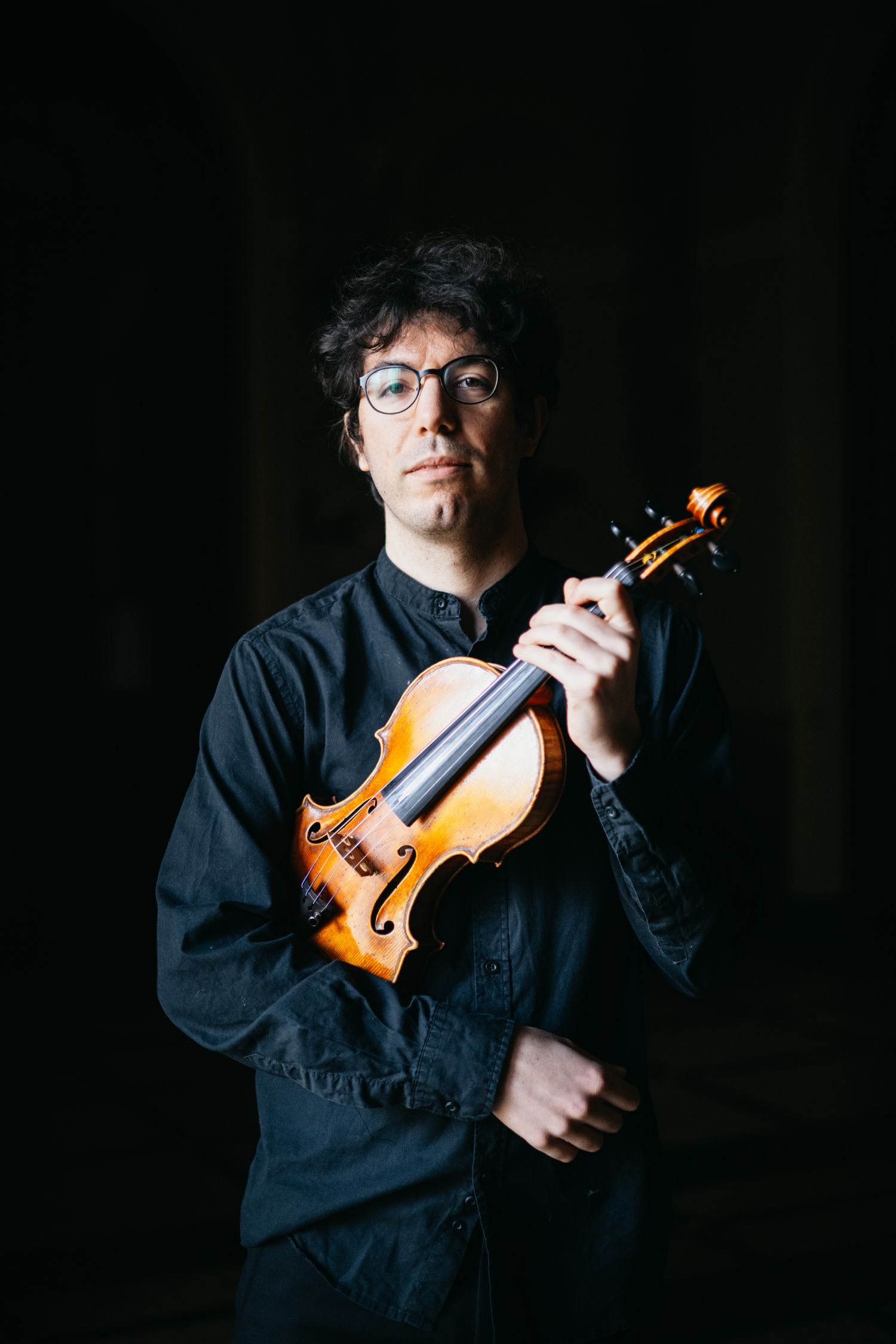 Male violinist wearing glasses poses with violin in dramatic dark lighting.
