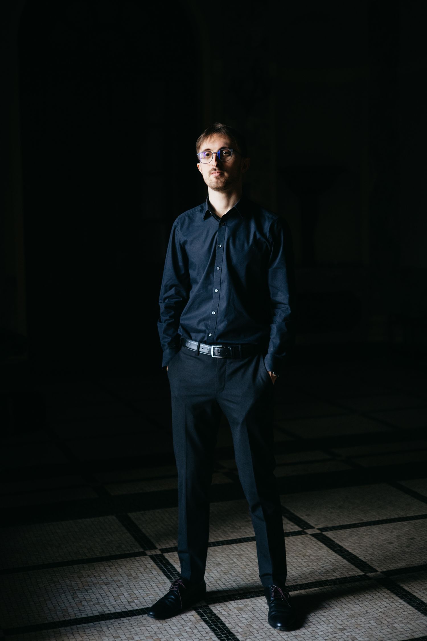 Young man in dark shirt and trousers posing with hands in pockets in dramatic lighting.