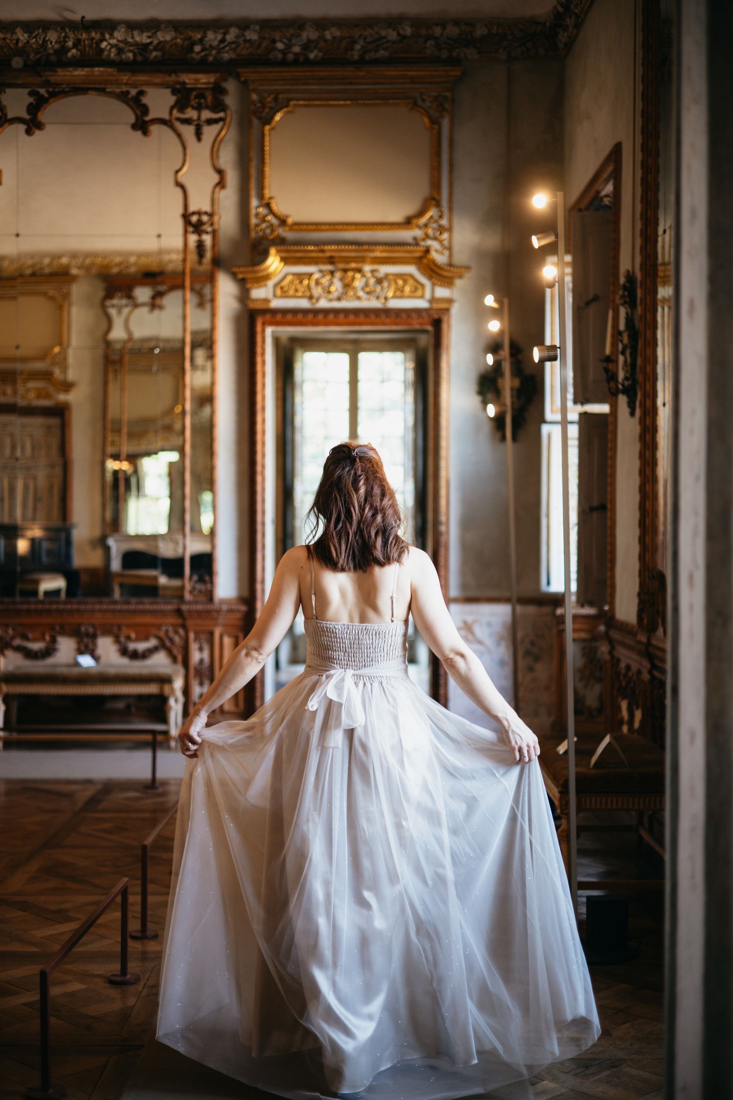 Bride in white tulle gown viewed from behind in ornate gilded ballroom with antique mirrors and wood paneling.