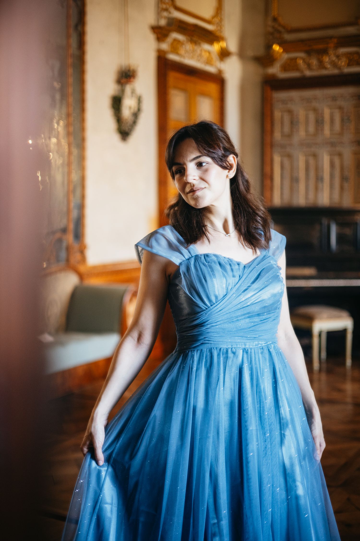 Woman in elegant blue tulle ball gown poses in ornate room with grand piano and gold decor.