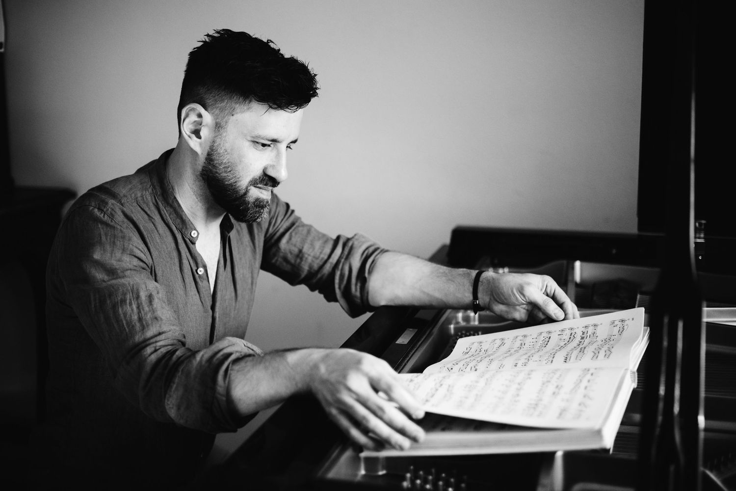 Black and white photo of bearded musician reviewing sheet music at a piano.