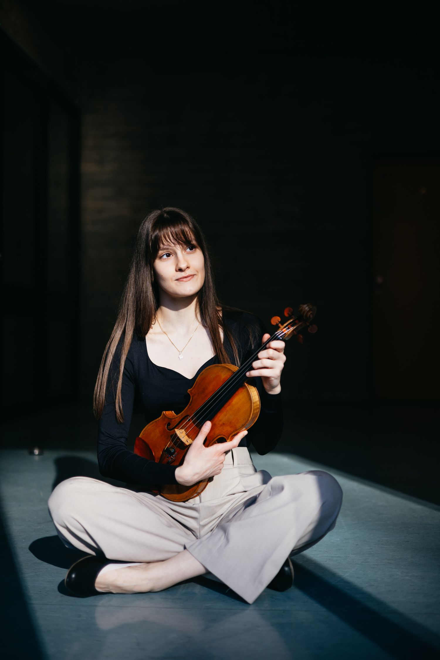 Young female musician sits cross-legged on floor holding a violin in a darkened studio space.