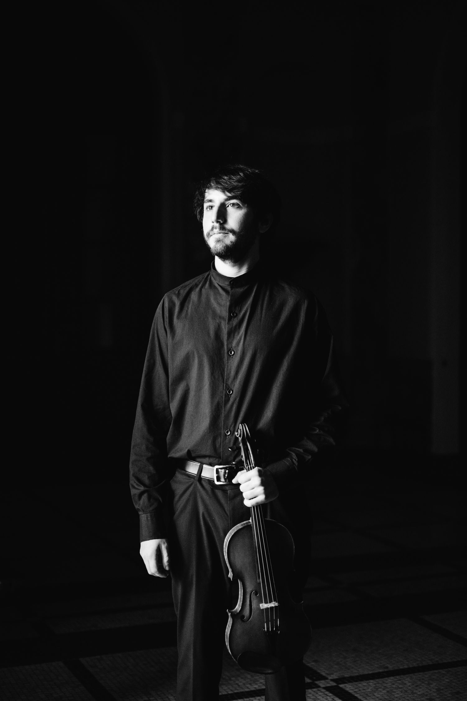 Black and white portrait of young male violinist holding instrument in dramatic low-key lighting.