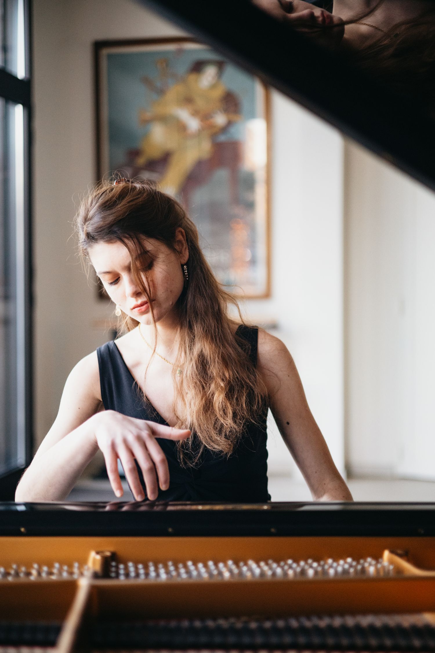 Young woman in black dress playing grand piano in elegant room with vintage poster on wall.