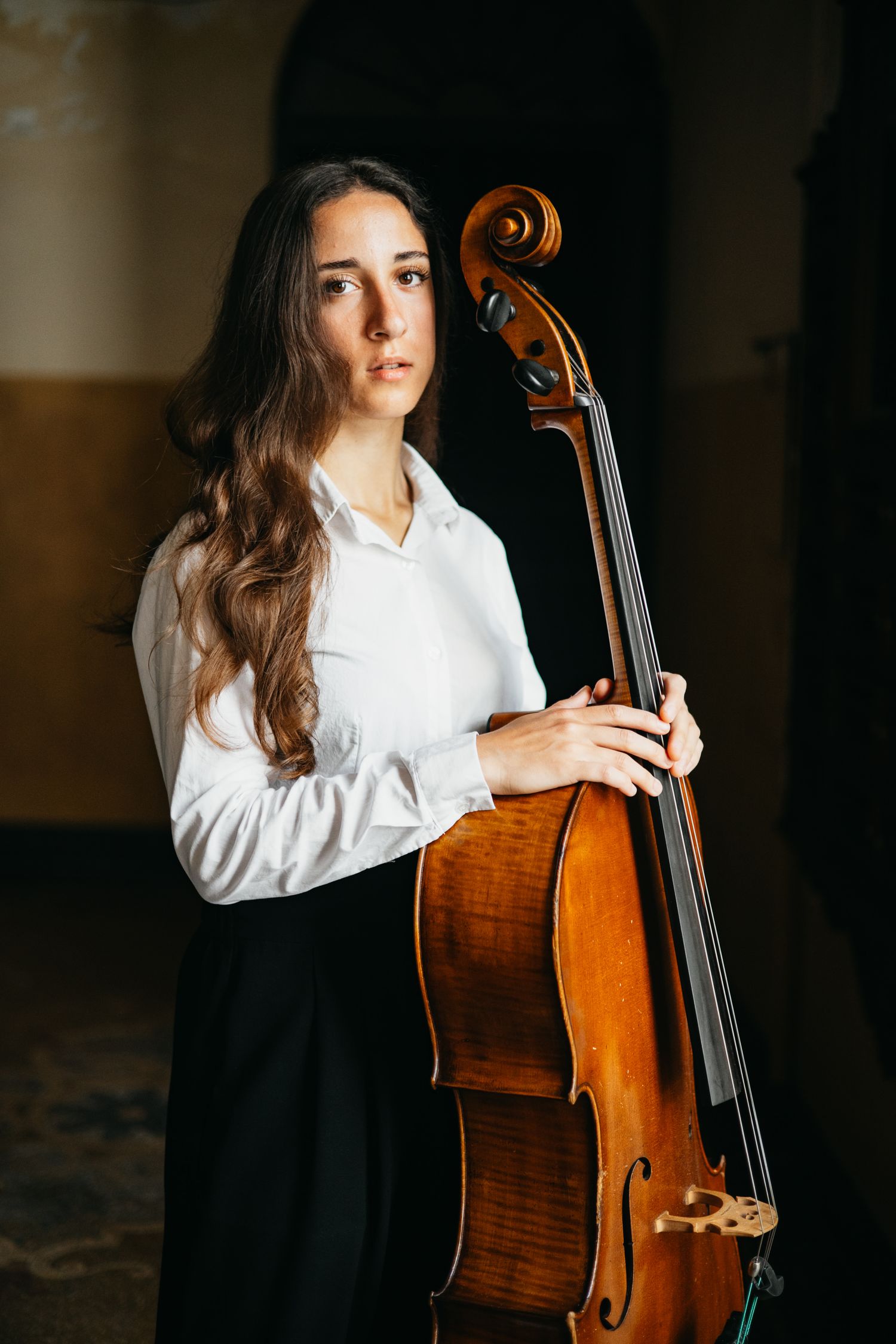 Young female cellist in white shirt holding cello upright in dramatic dark interior space.