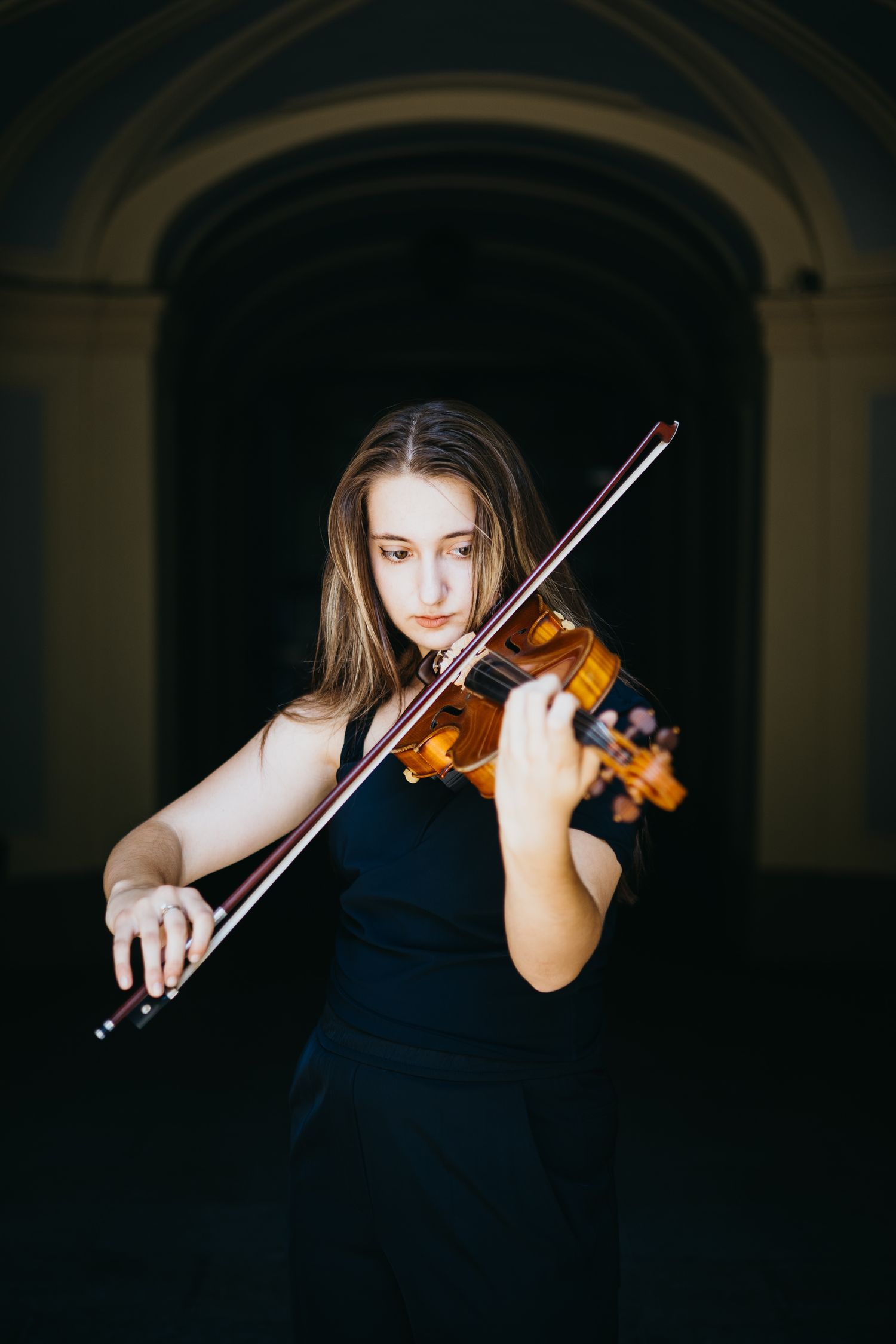 Young woman in black dress playing violin passionately under a dark arched corridor.