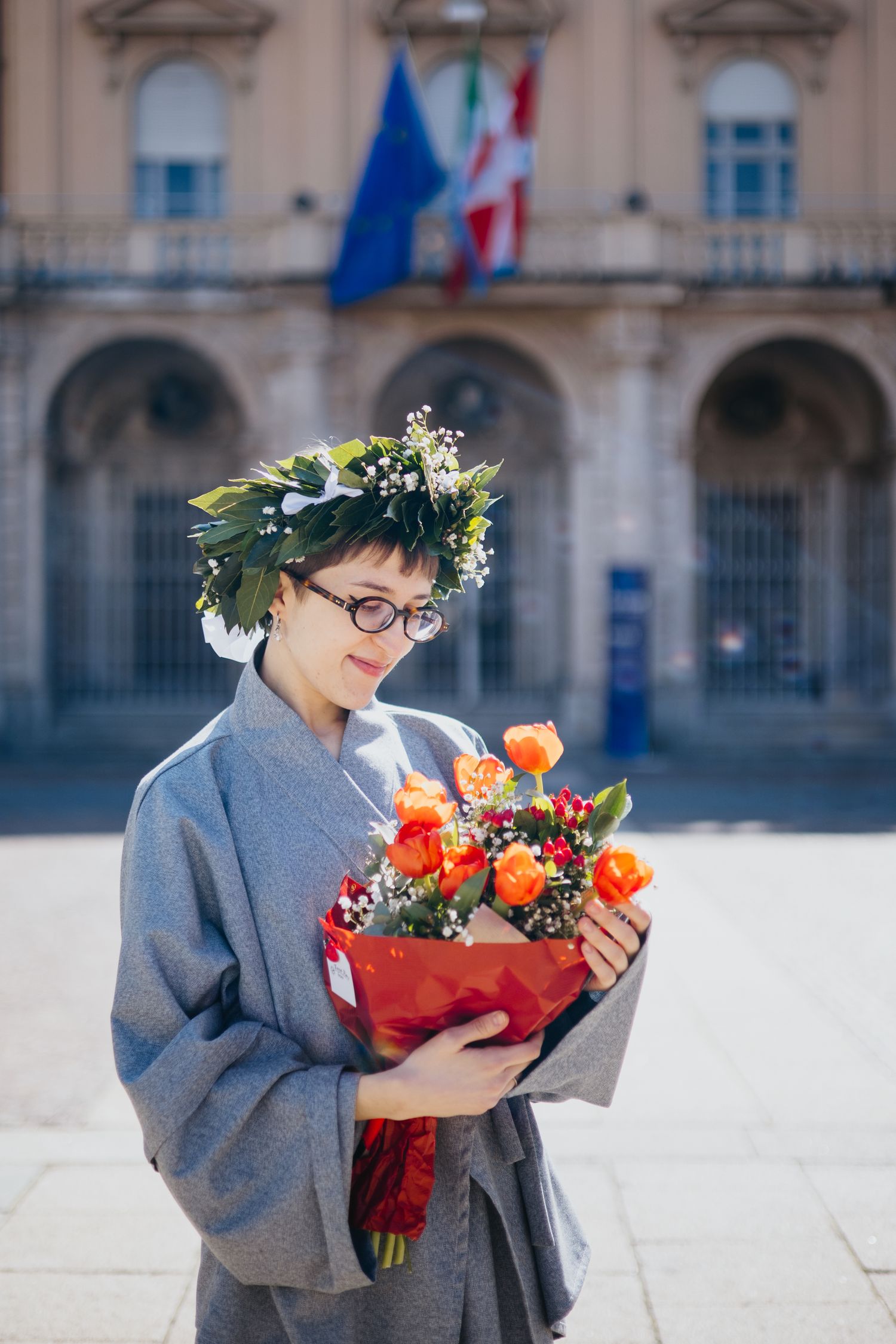 Young woman in graduation robe and laurel wreath admires orange rose bouquet in Italian piazza.