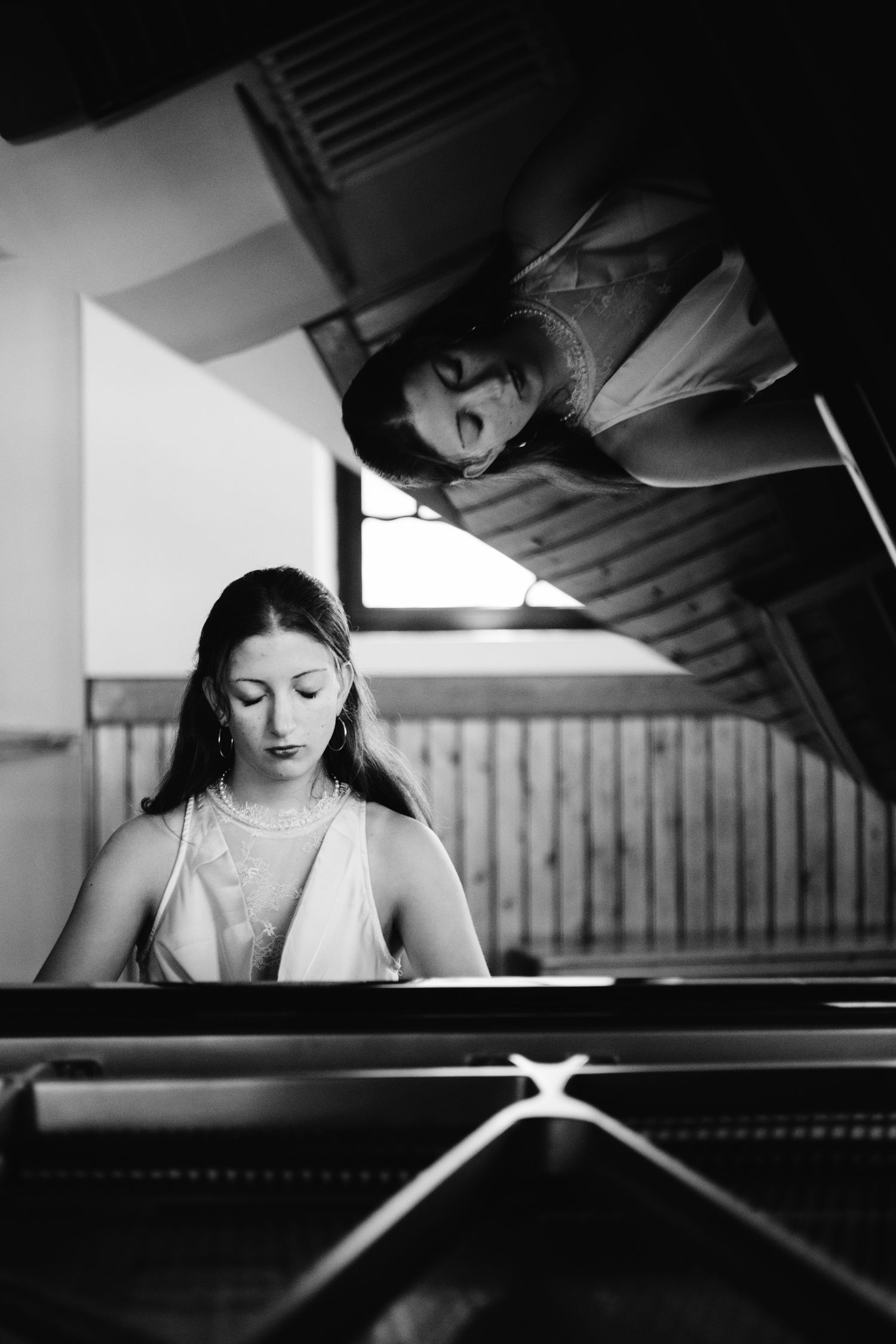 Black and white photo of a young woman playing grand piano, her reflection visible in the raised lid above.