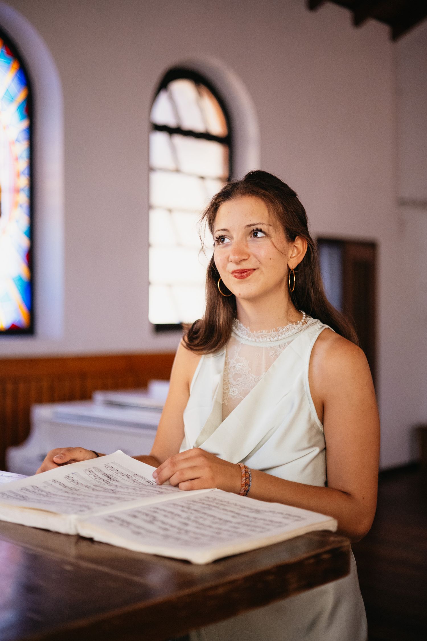 Young woman in white dress reading sheet music at a church podium with stained glass windows behind her.