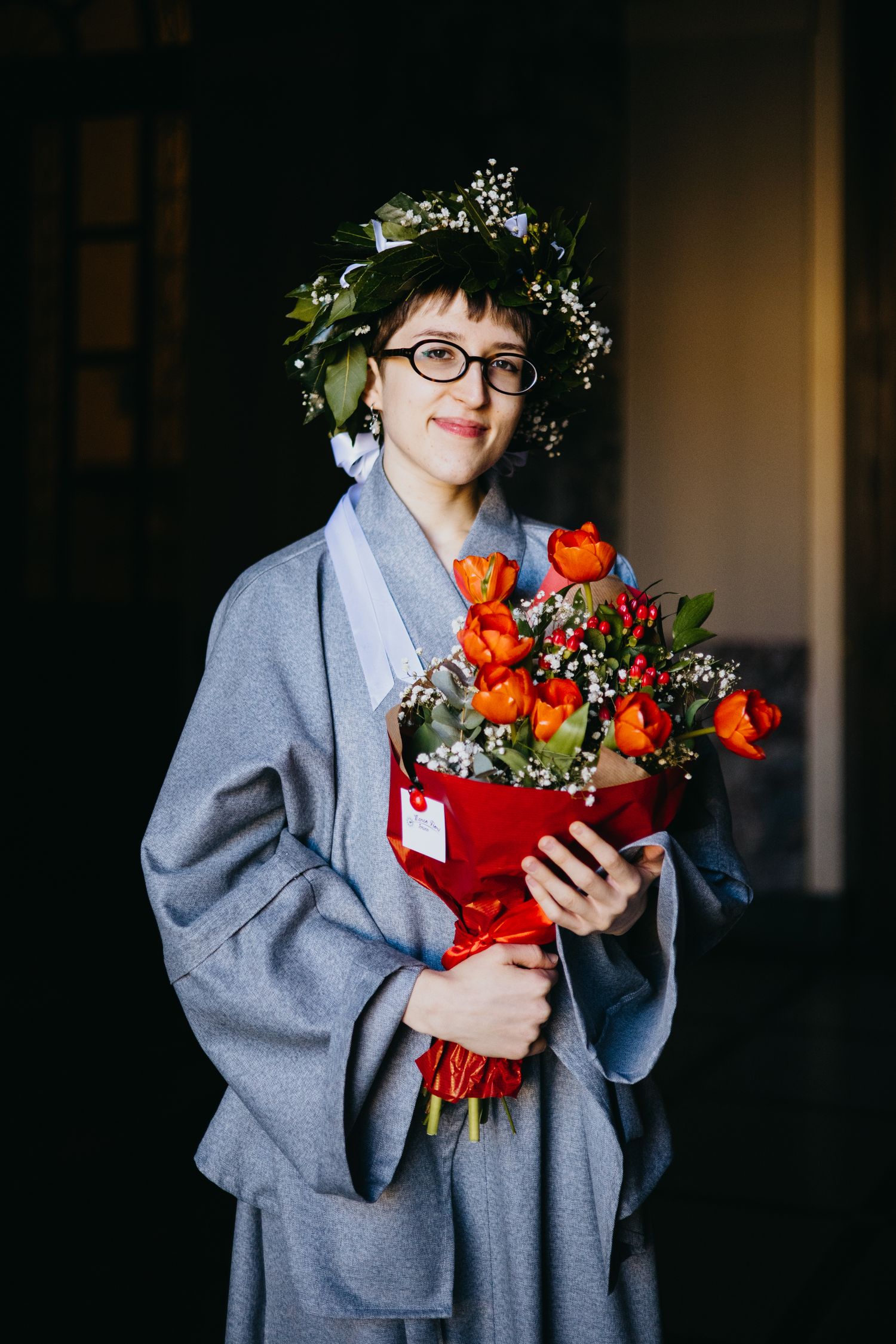 Woman in gray coat holding red tulip bouquet wearing floral crown and glasses.