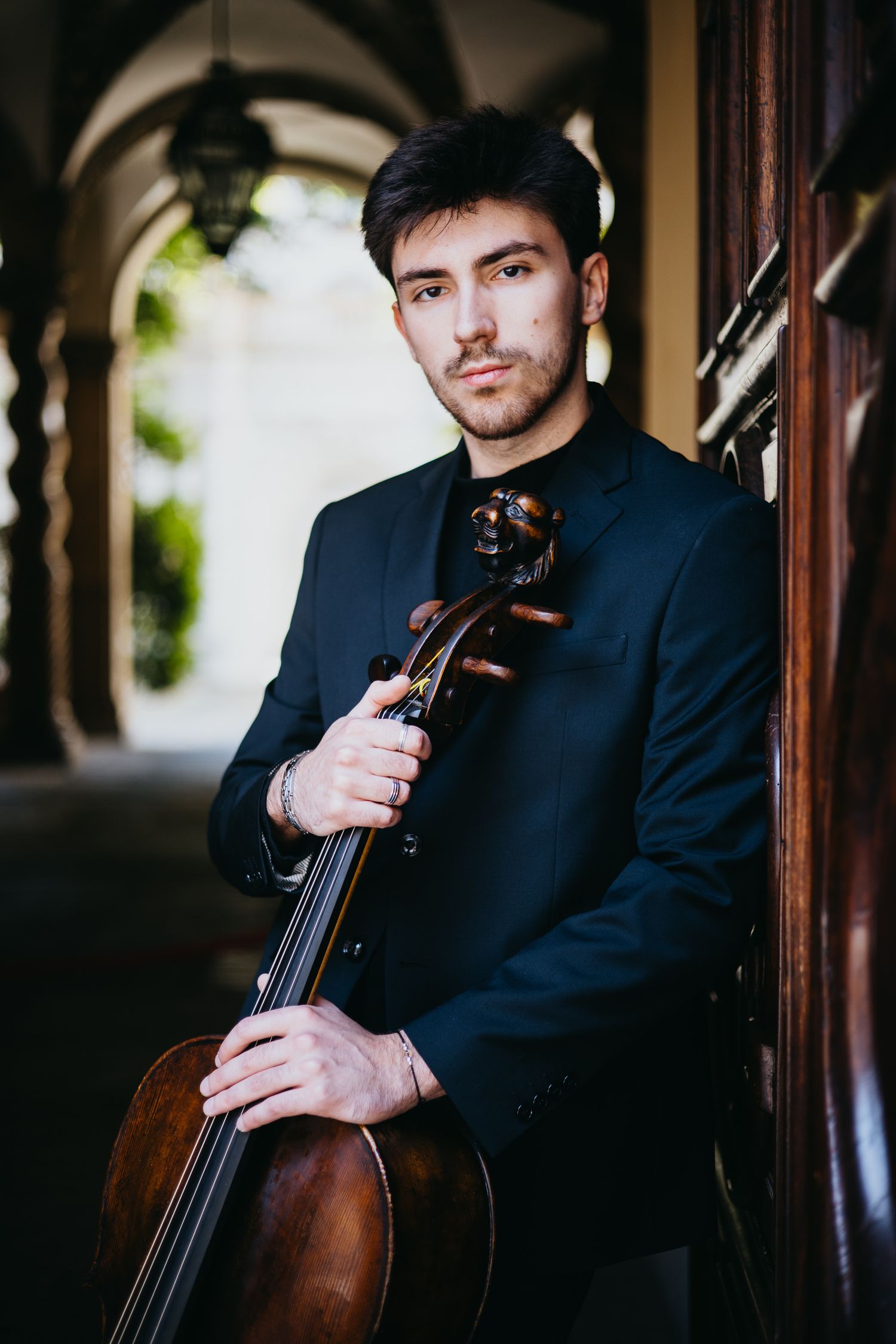 Young male cellist in black suit leans against a wooden door holding his cello in a classical architectural setting.