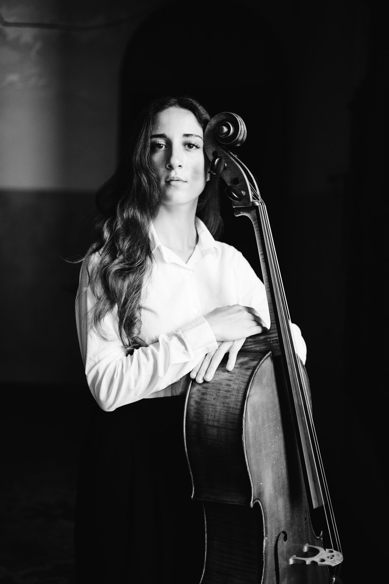Black and white portrait of a young female cellist holding her cello in a dimly lit room.