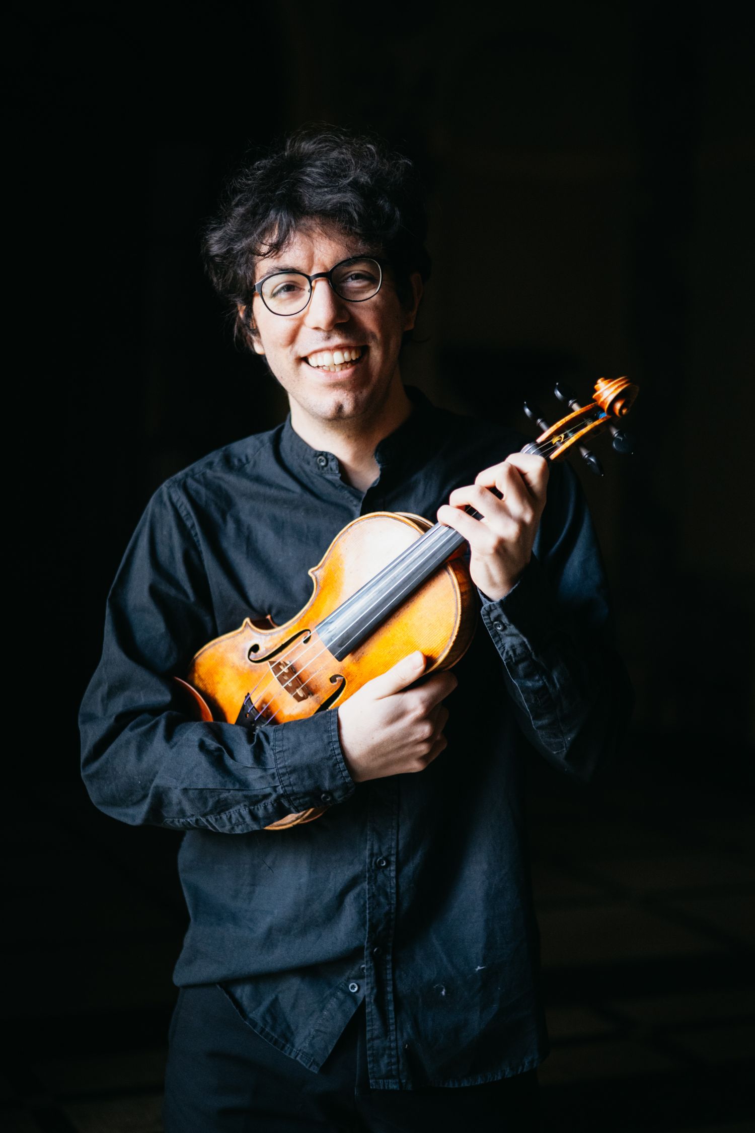 Smiling violinist with glasses holds violin in dark atmospheric setting.