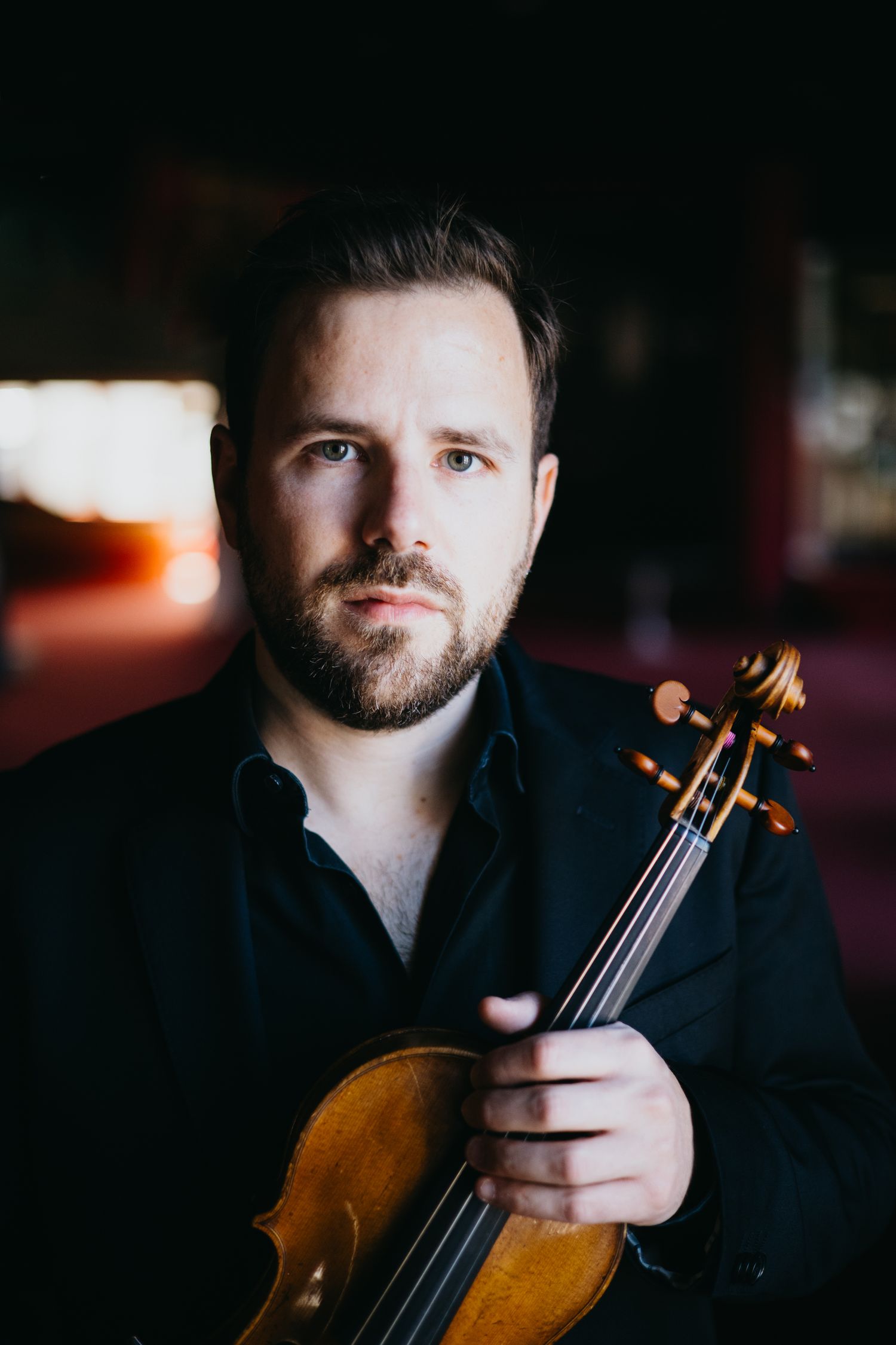 A bearded male violinist in a black shirt holds his violin, posing for a professional portrait in dramatic lighting.