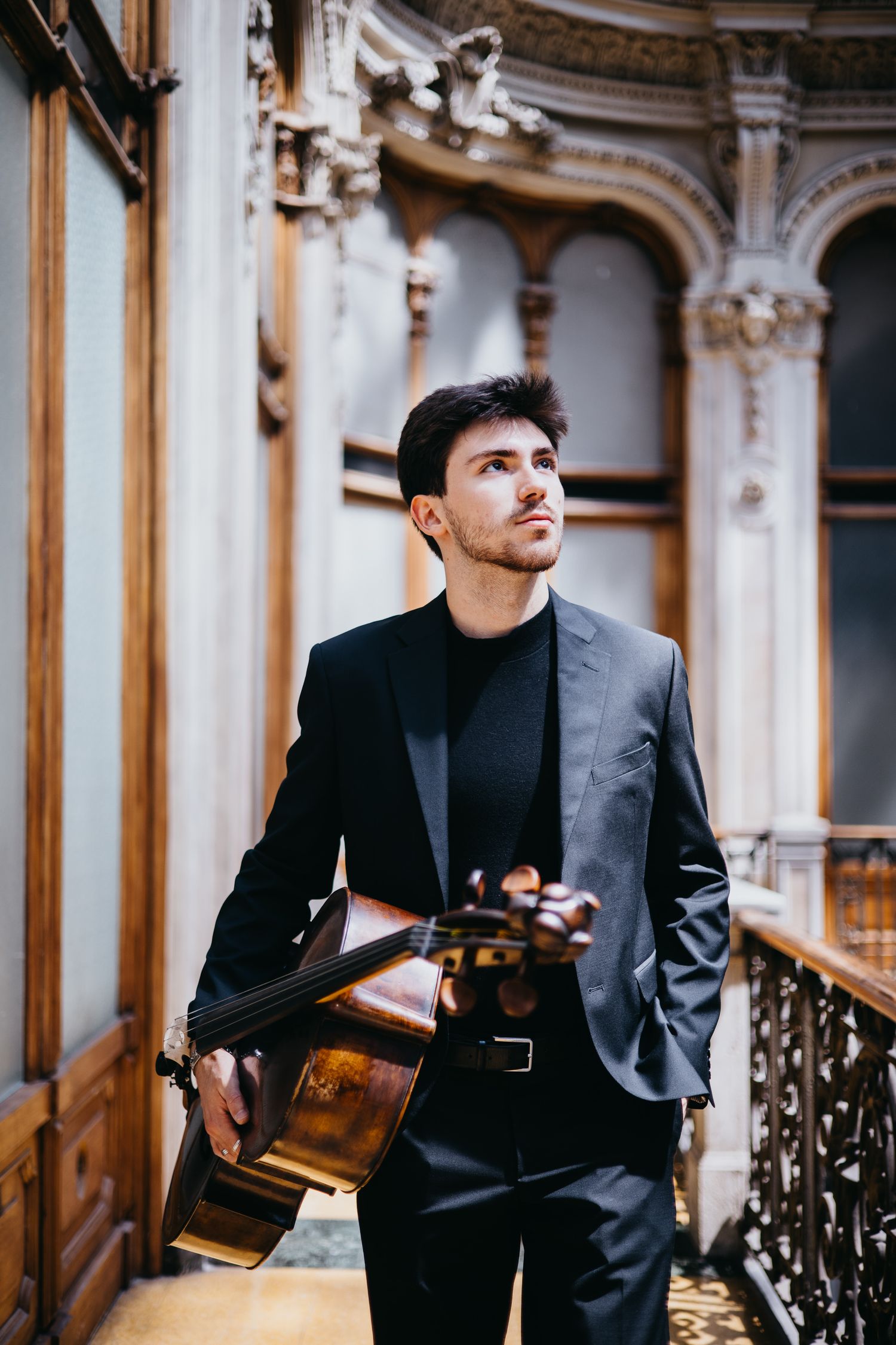 Elegant male cellist holding cello in ornate baroque building interior wearing black suit.