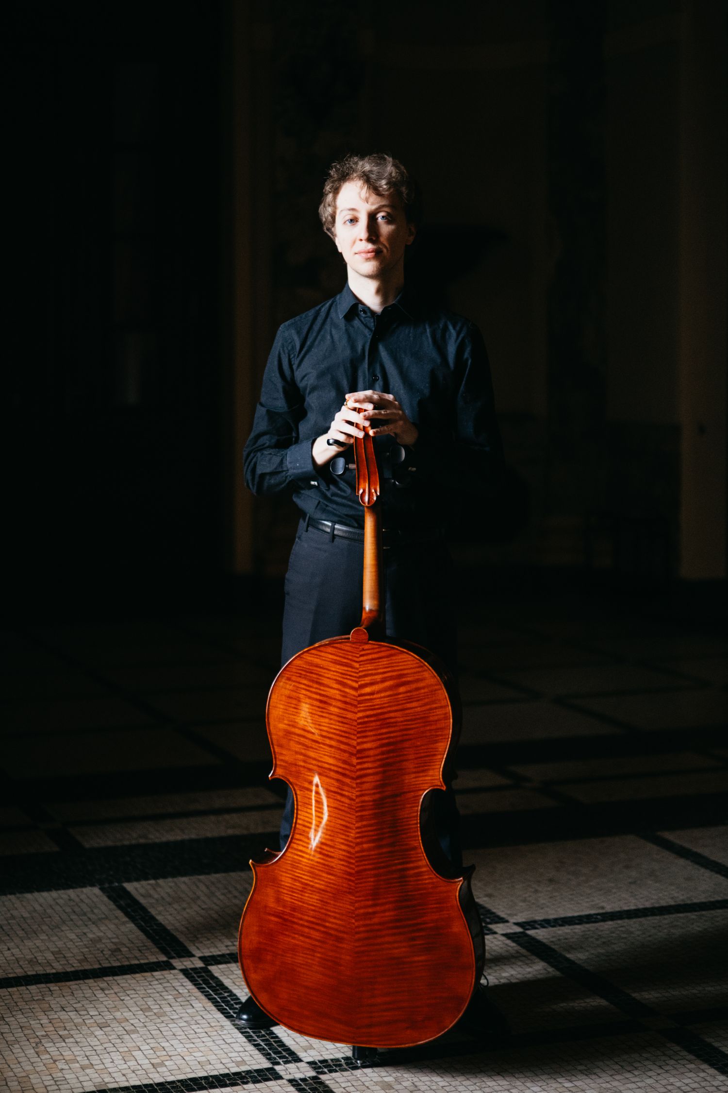 Cellist displays back of cello in dramatic dark architectural setting.