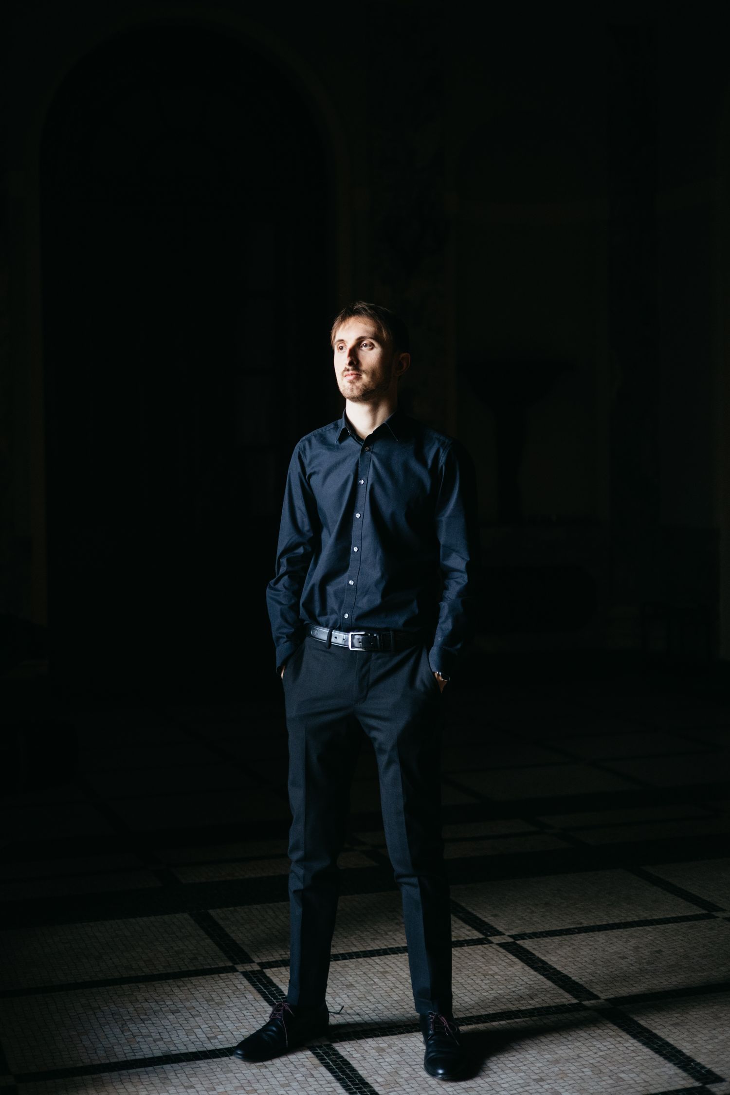 Young man in navy blue shirt and trousers standing confidently in dramatic dark setting.