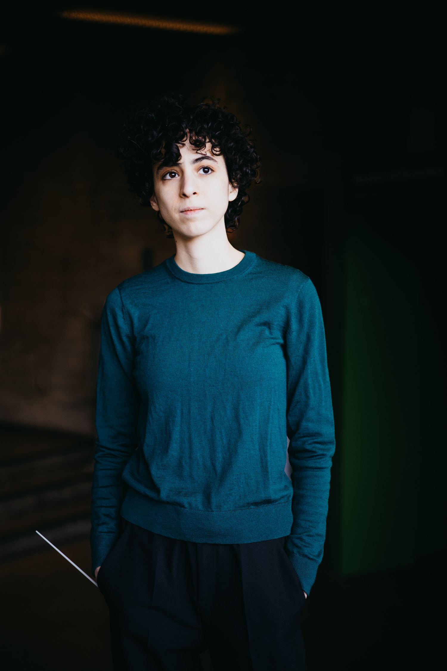 Young conductor in teal sweater holding baton in dark hallway.