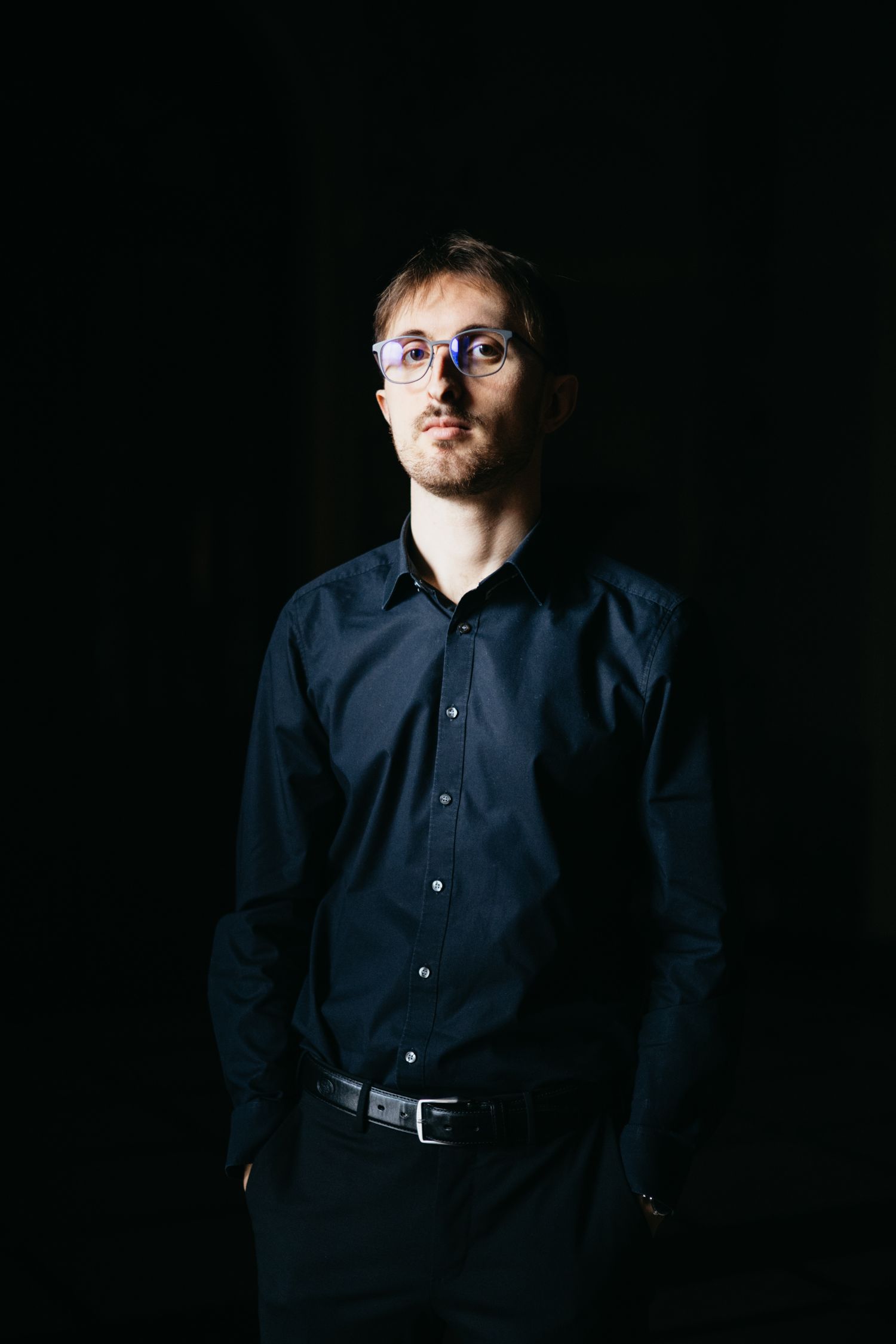 Young man wearing glasses in dark blue shirt posing against black background in studio portrait.