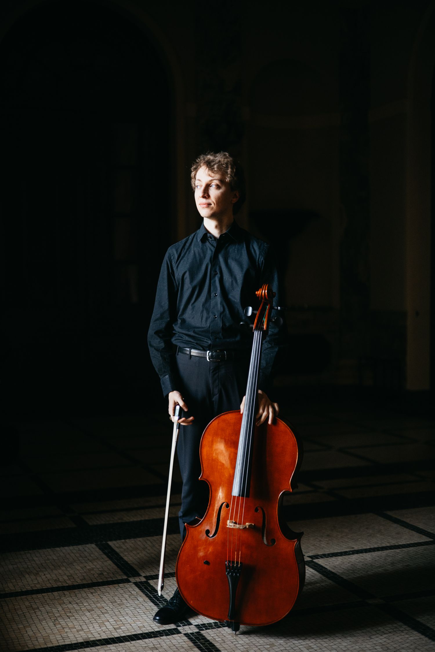 Young cellist in black attire stands with cello and bow on tiled floor.