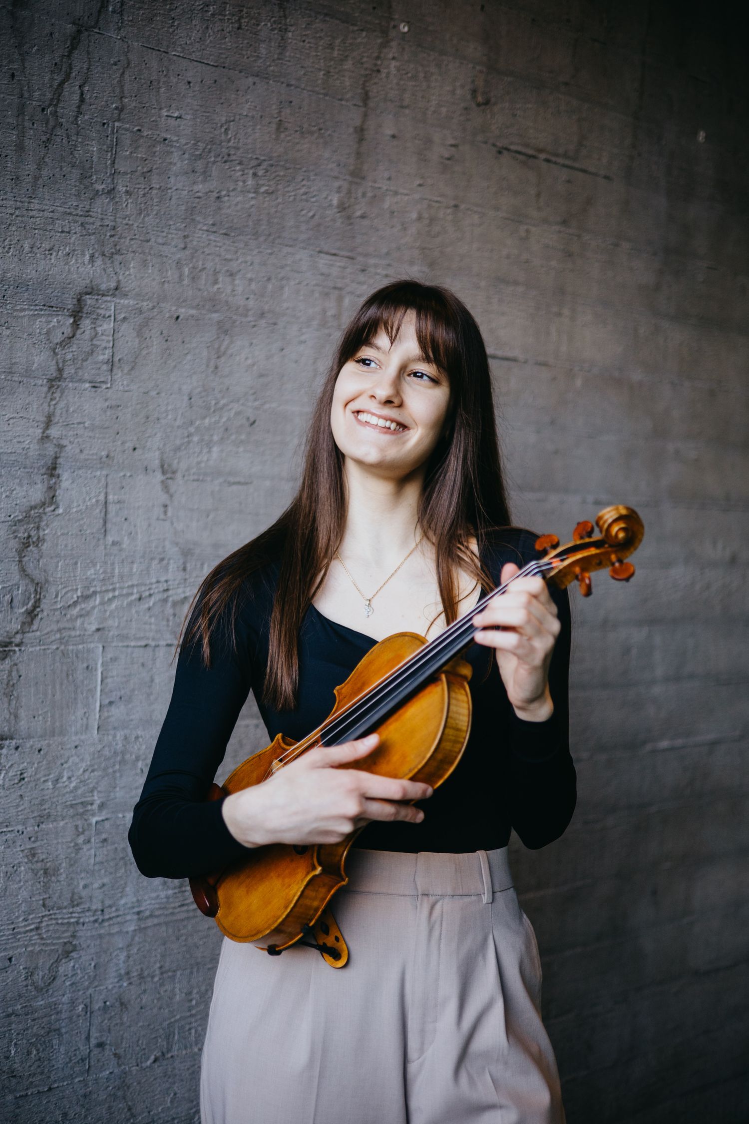 Smiling young female violinist holding a violin against a gray concrete wall.