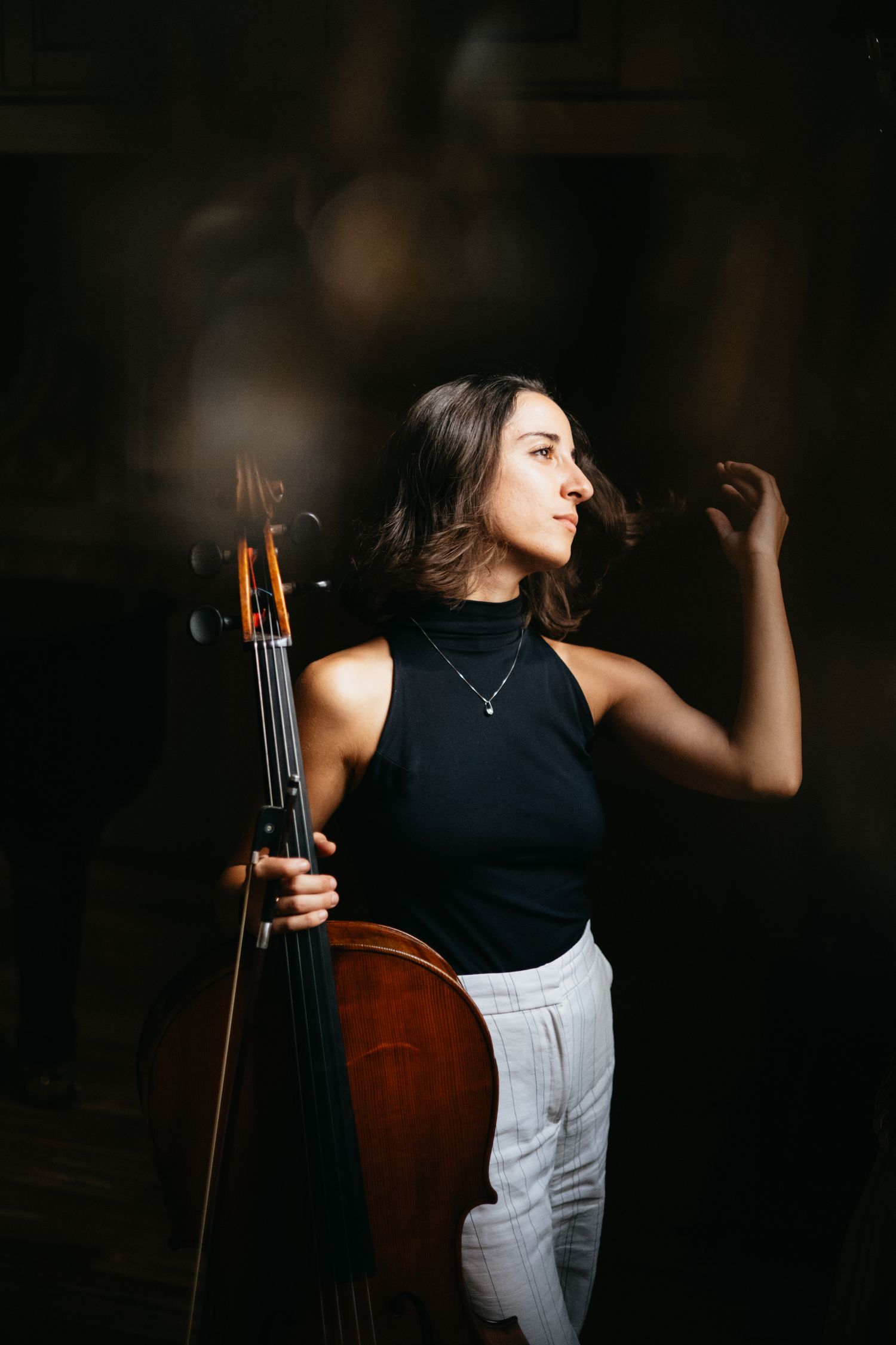 A young woman in a black sleeveless top holds a cello against a dark background, gazing thoughtfully to the side.