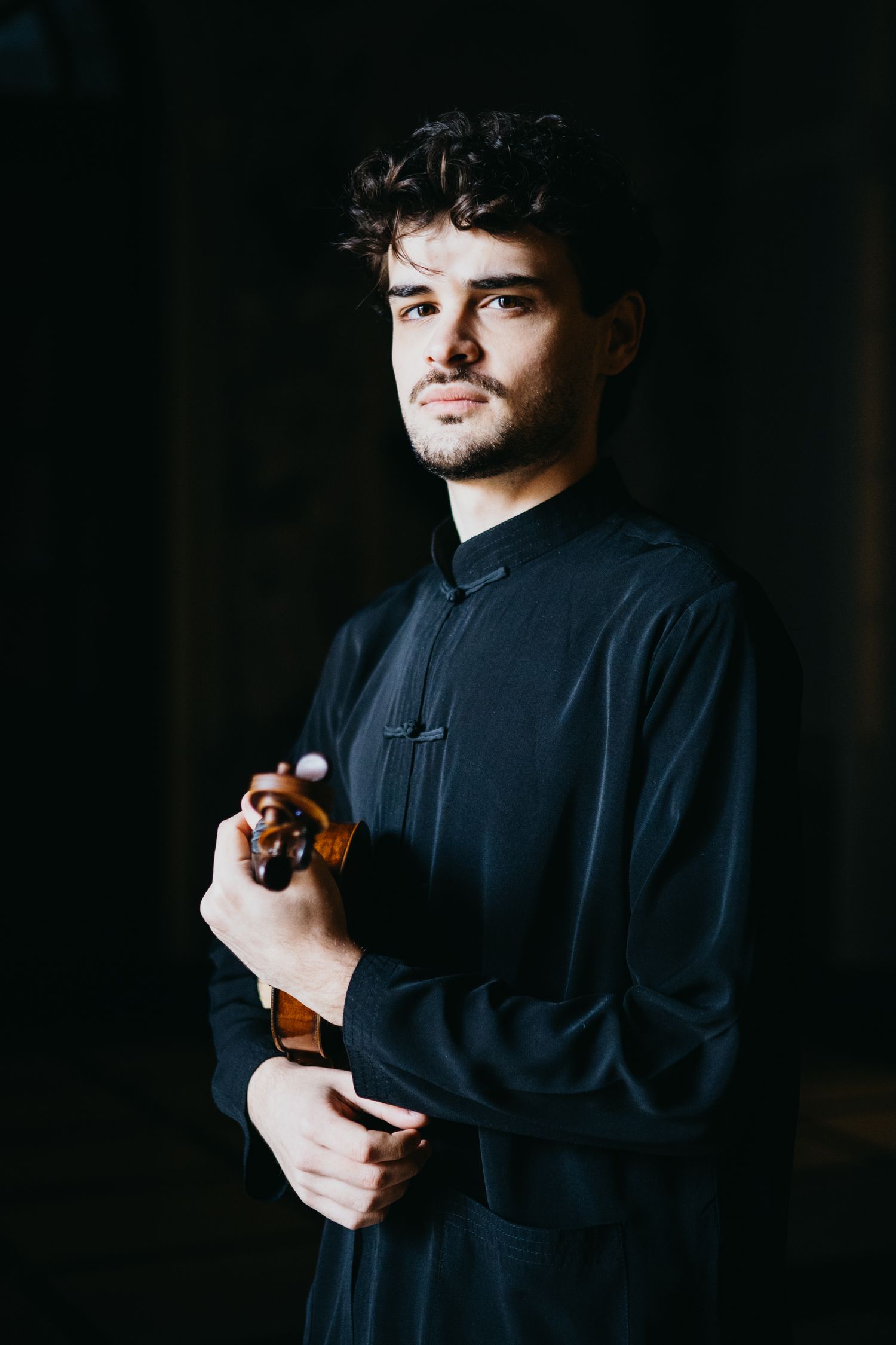Young male musician in black shirt holds violin in dramatic low-key lighting.