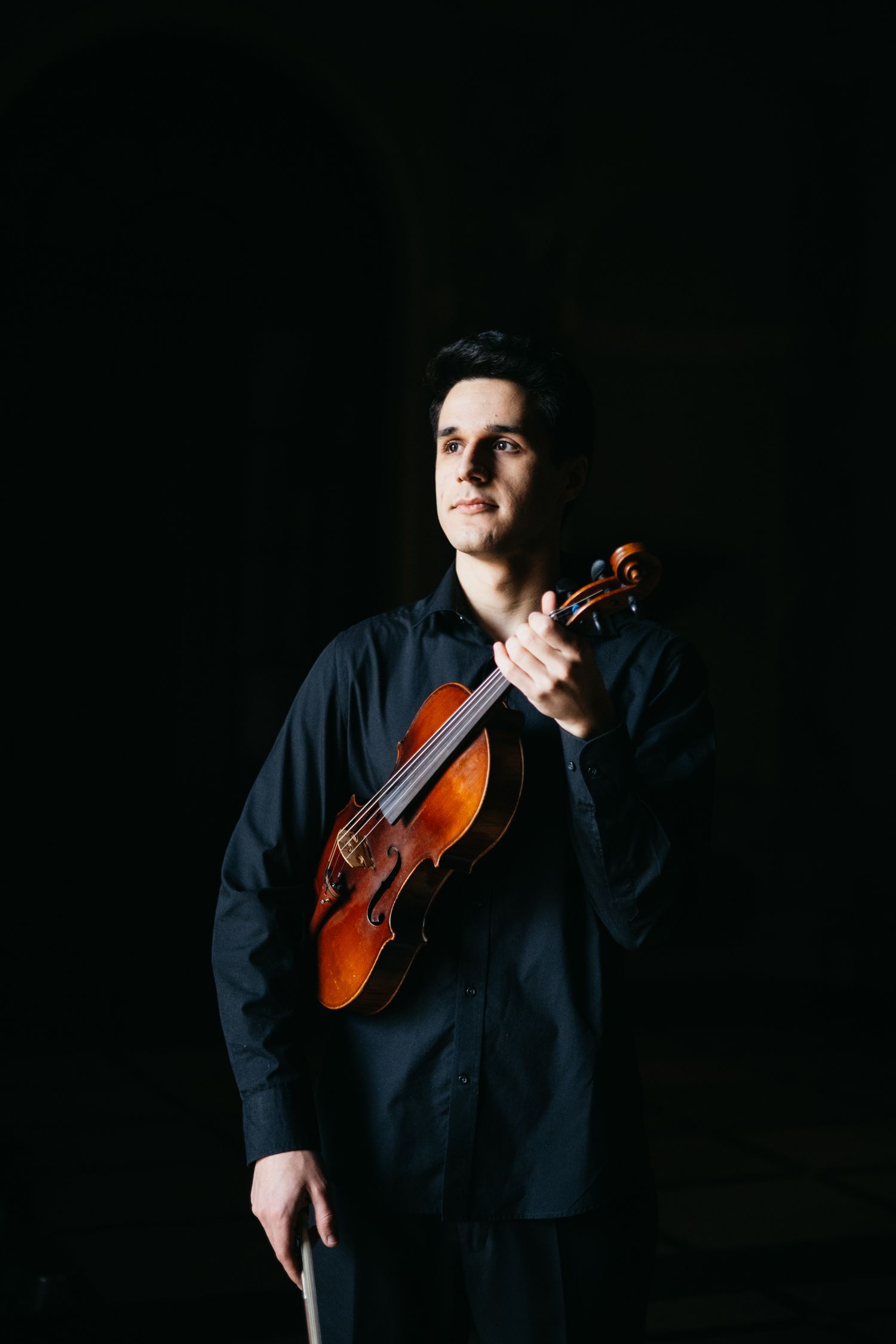Male violinist in black shirt holds violin upward in dramatic low-key lighting portrait.