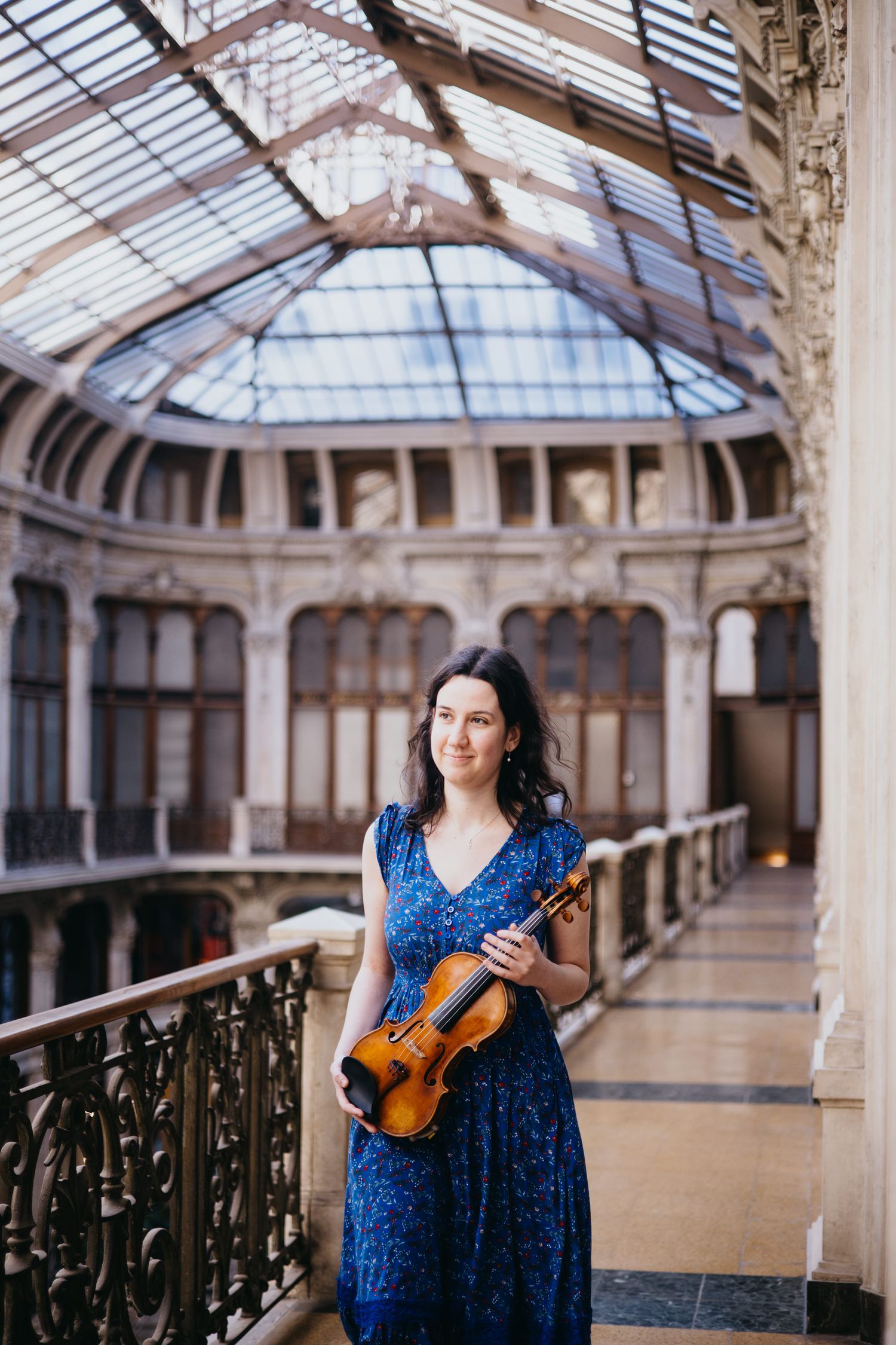 Woman in blue dress holding a violin stands on a balcony inside an ornate historic building with glass ceiling.