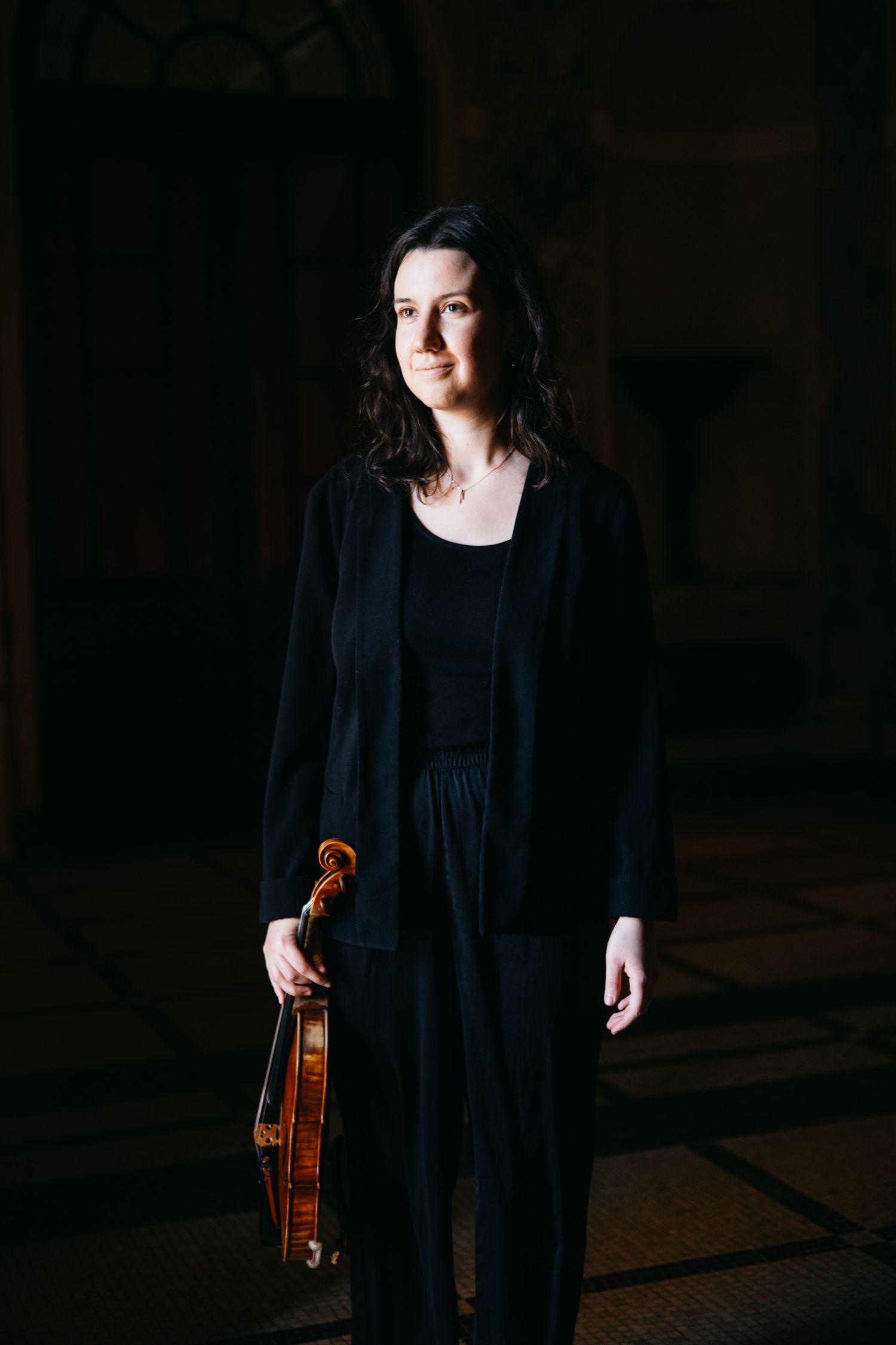 A female violinist in black attire holds her violin while standing in a dimly lit architectural space.