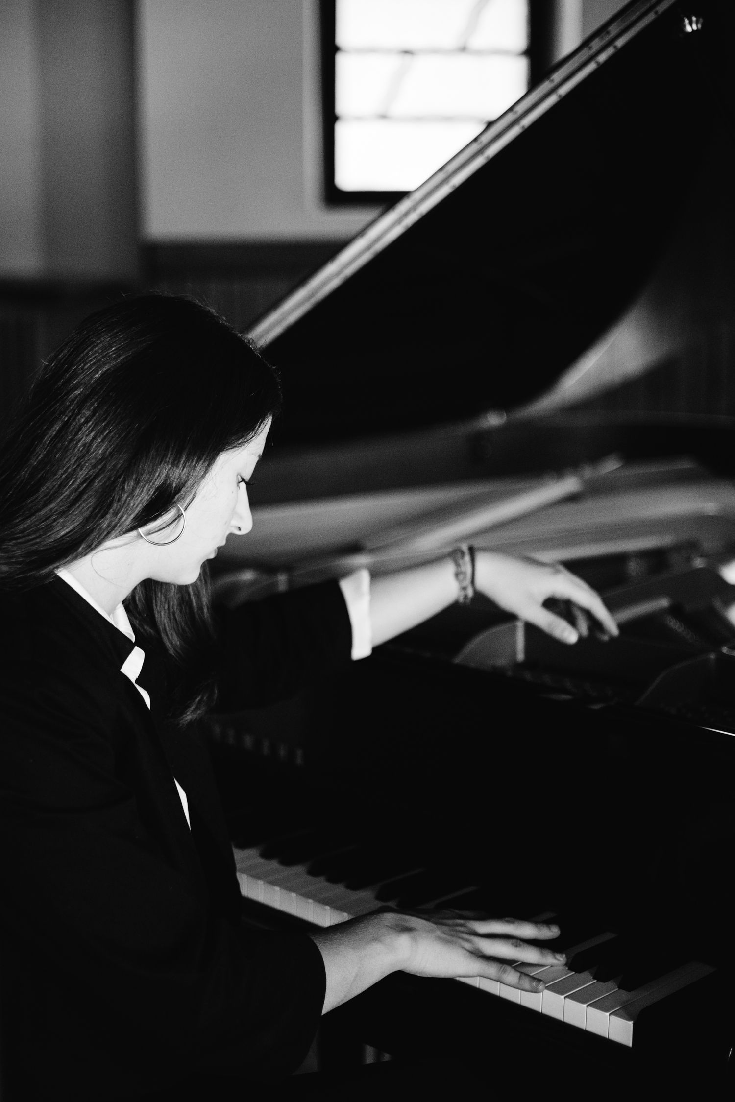 Black and white photo of a young woman with long hair playing a grand piano in a dimly lit room.