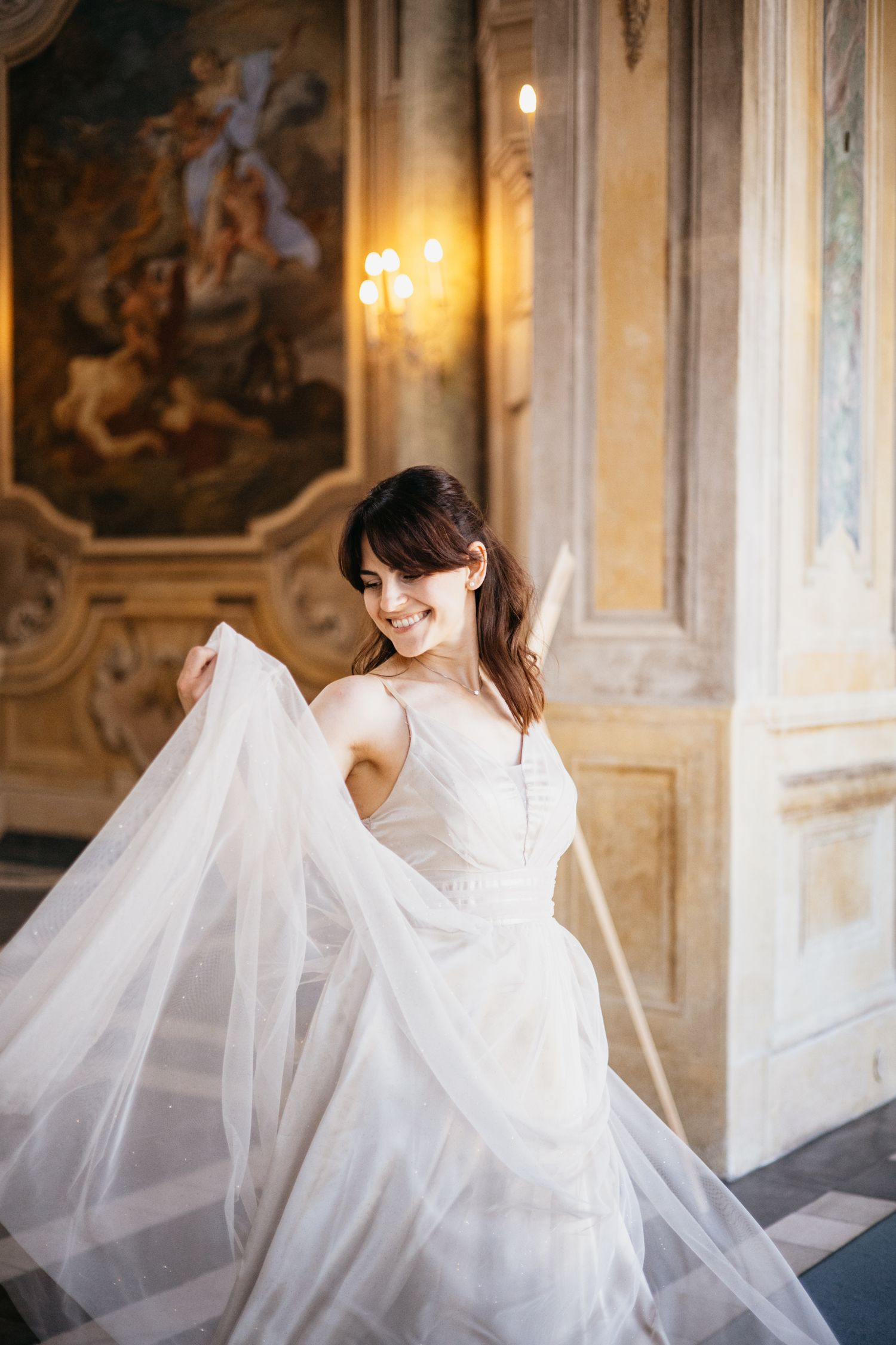 Smiling bride in white tulle gown twirling in ornate baroque hall with golden frescoes and candlelight.