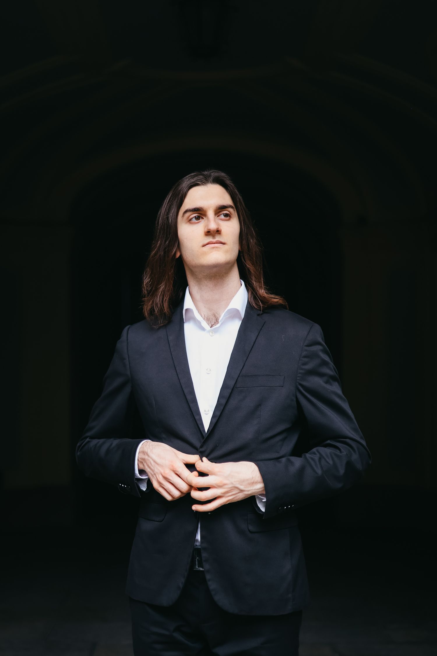 Young man with long dark hair in a dark blazer and white shirt, standing confidently in a dramatic dark archway.