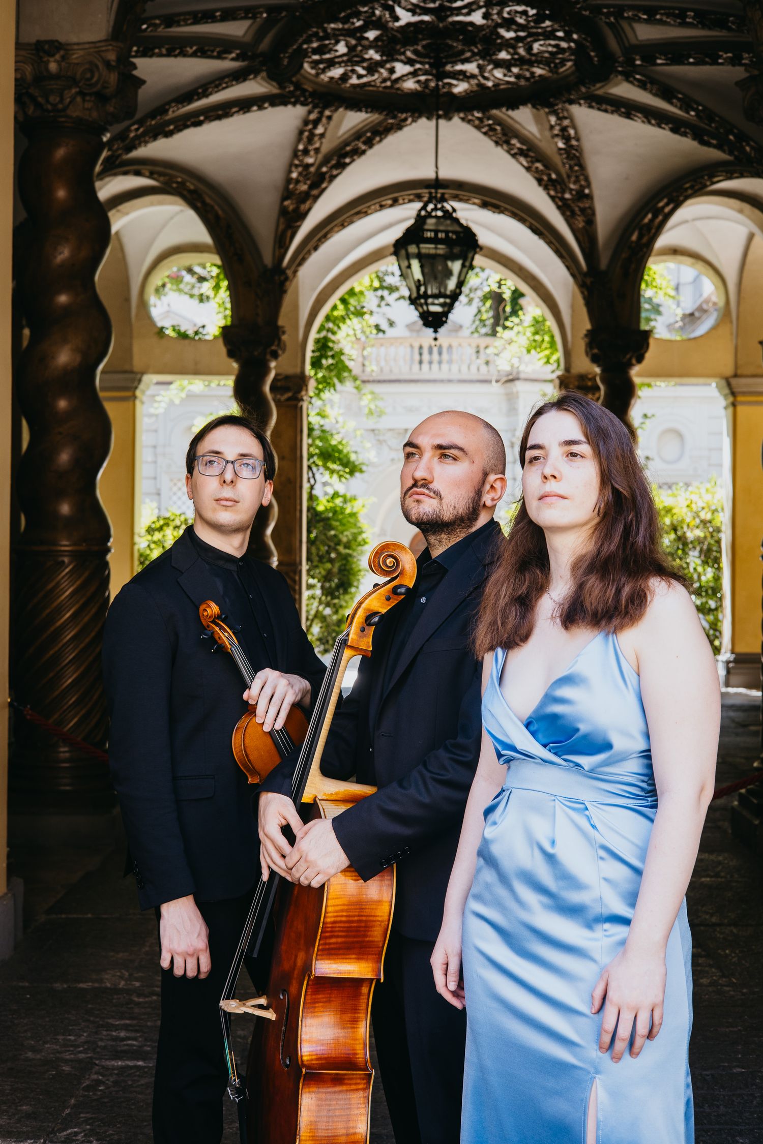 A classical music trio poses with cello and violin under elegant arched architecture in formal wear.
