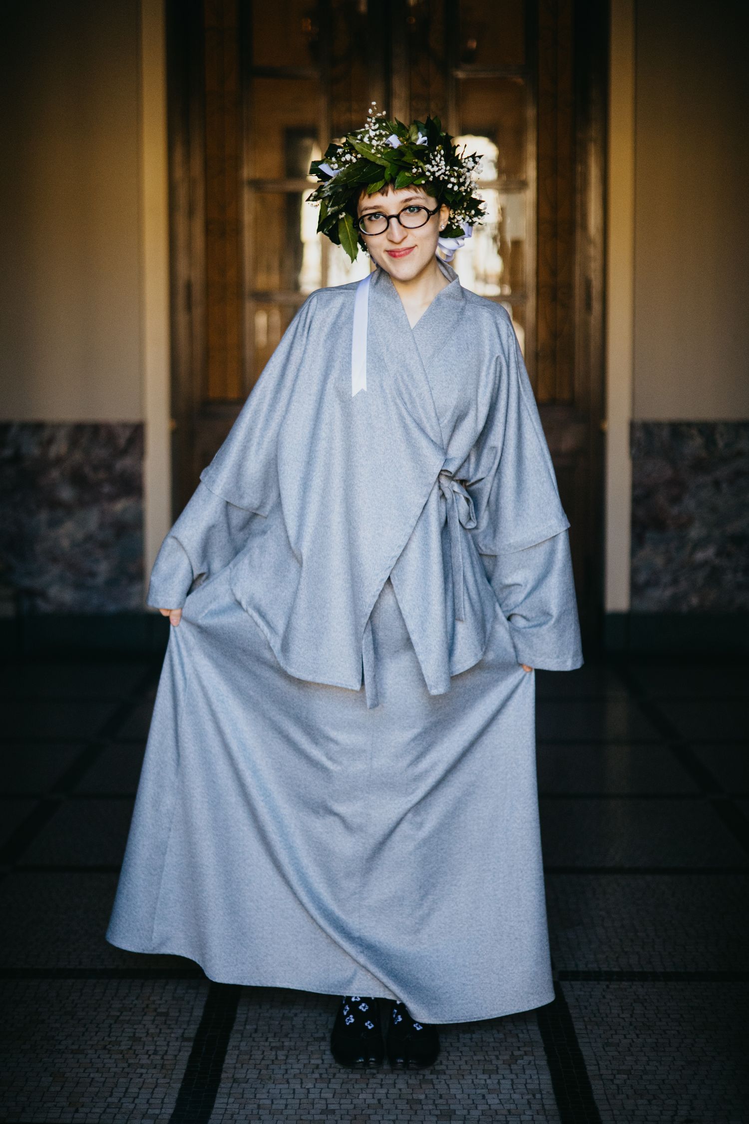 Smiling graduate poses in elegant gray academic robe and green floral crown in ornate hallway.