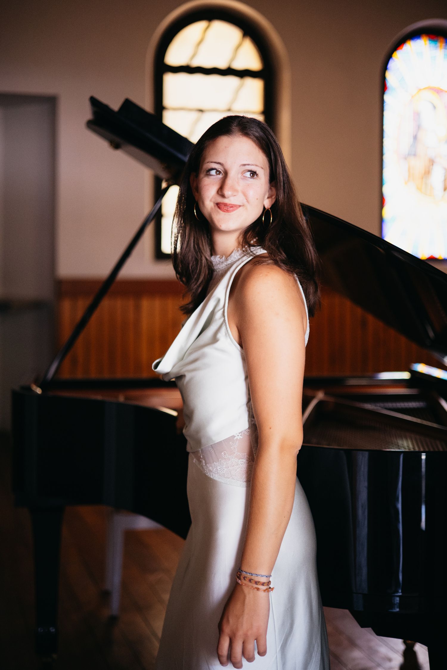 Woman in elegant silver dress standing by a grand piano in a church with stained glass windows.