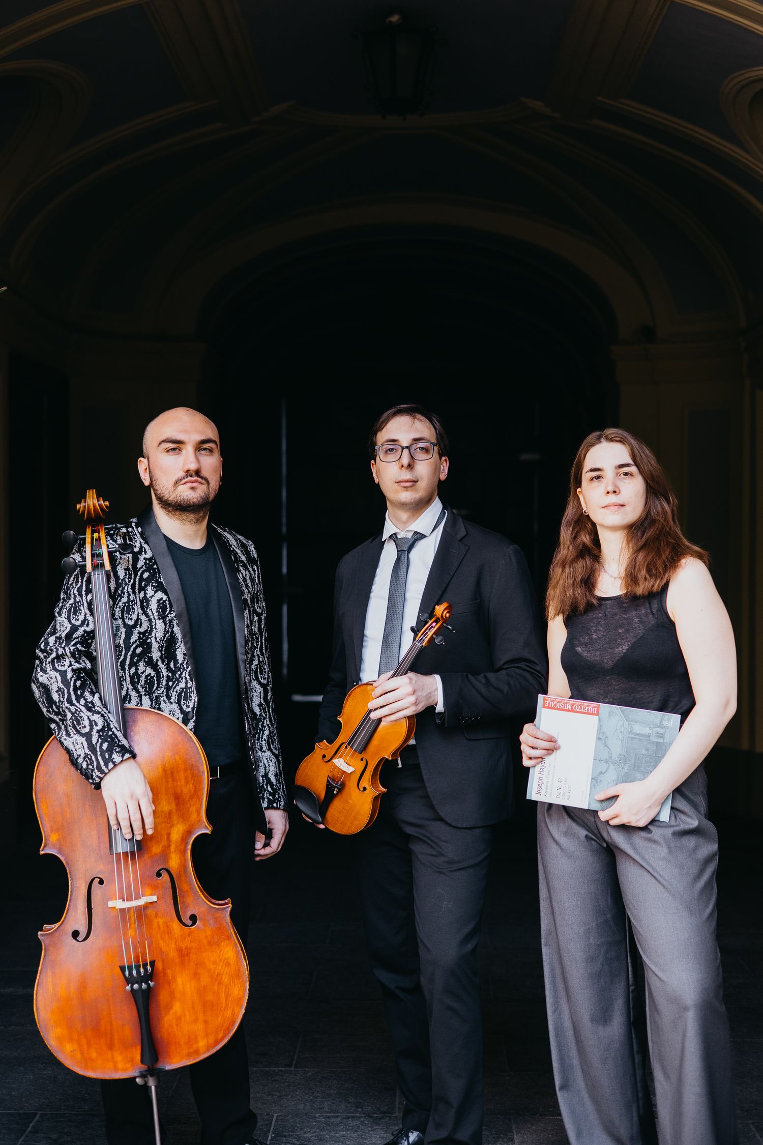 Classical music trio standing under arch with cello and violin instruments outdoors.