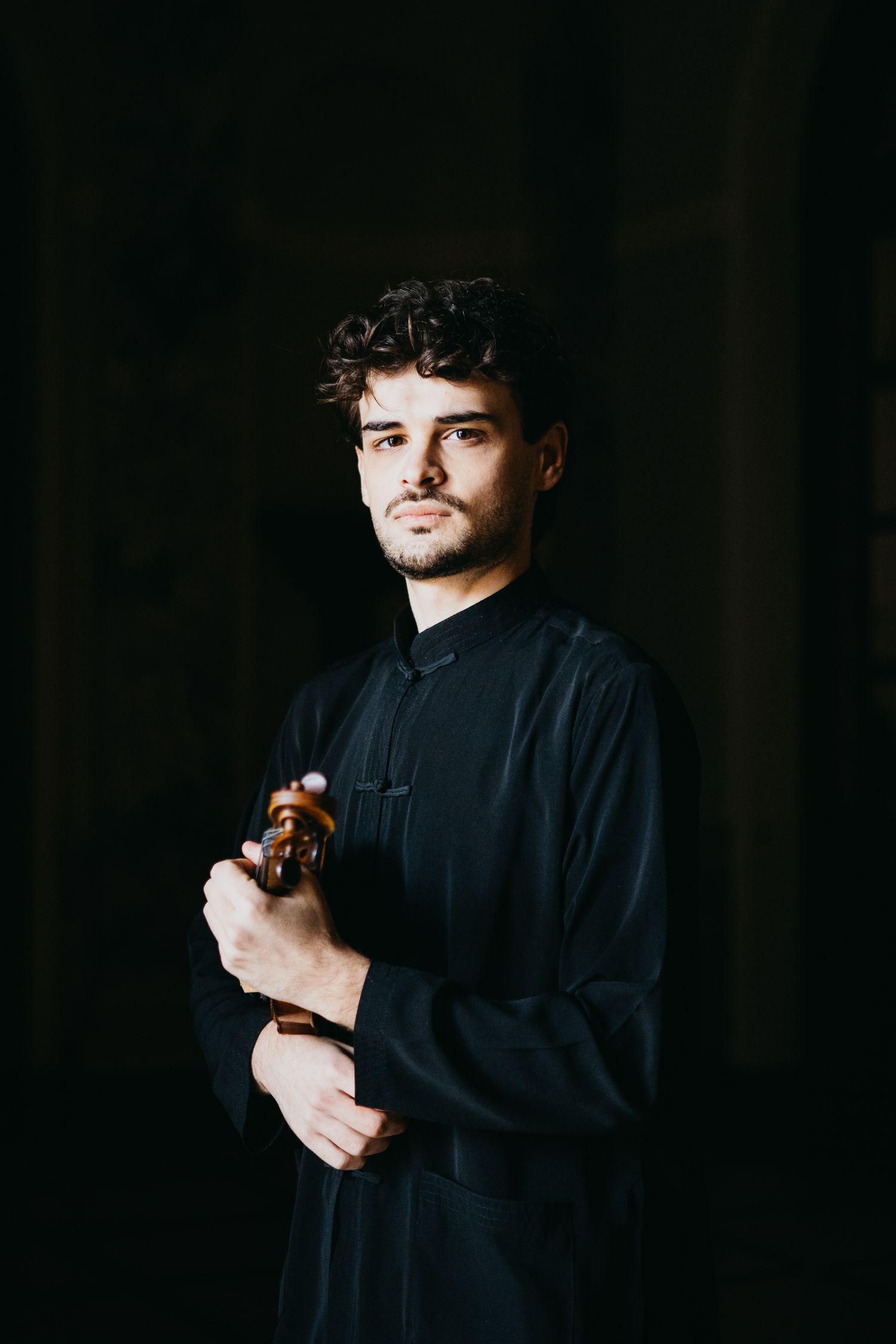 Male violinist in black outfit cradles violin against dark moody background.