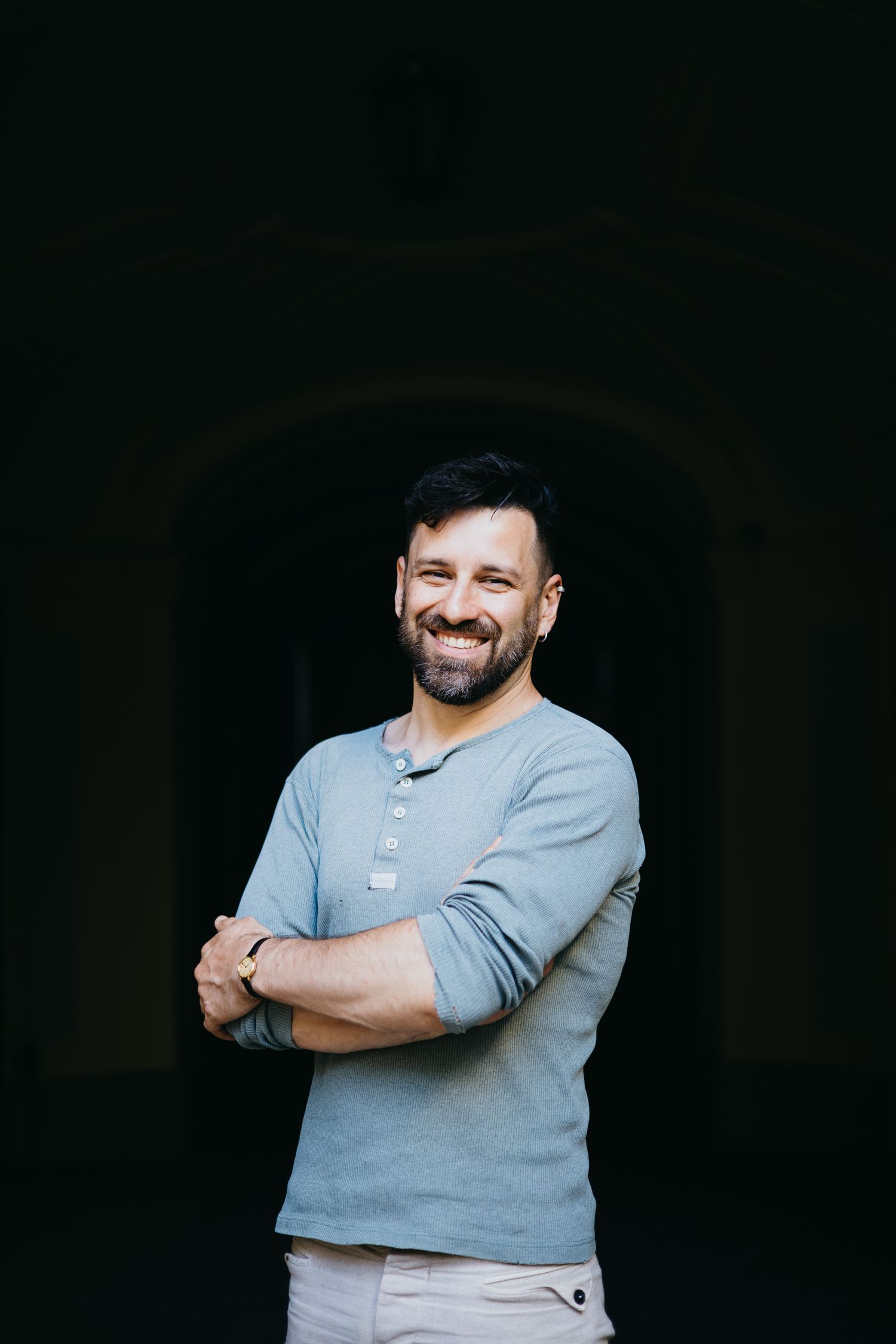 Cheerful bearded man in gray shirt smiling with arms crossed against dark archway background.