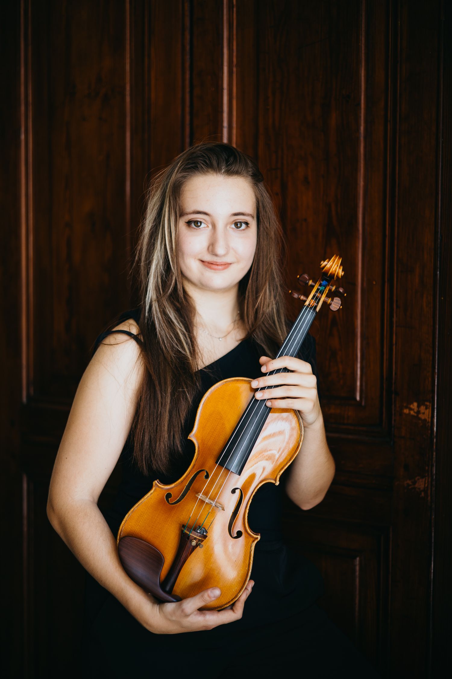 Smiling young female violinist in black dress holds violin against wooden door background.