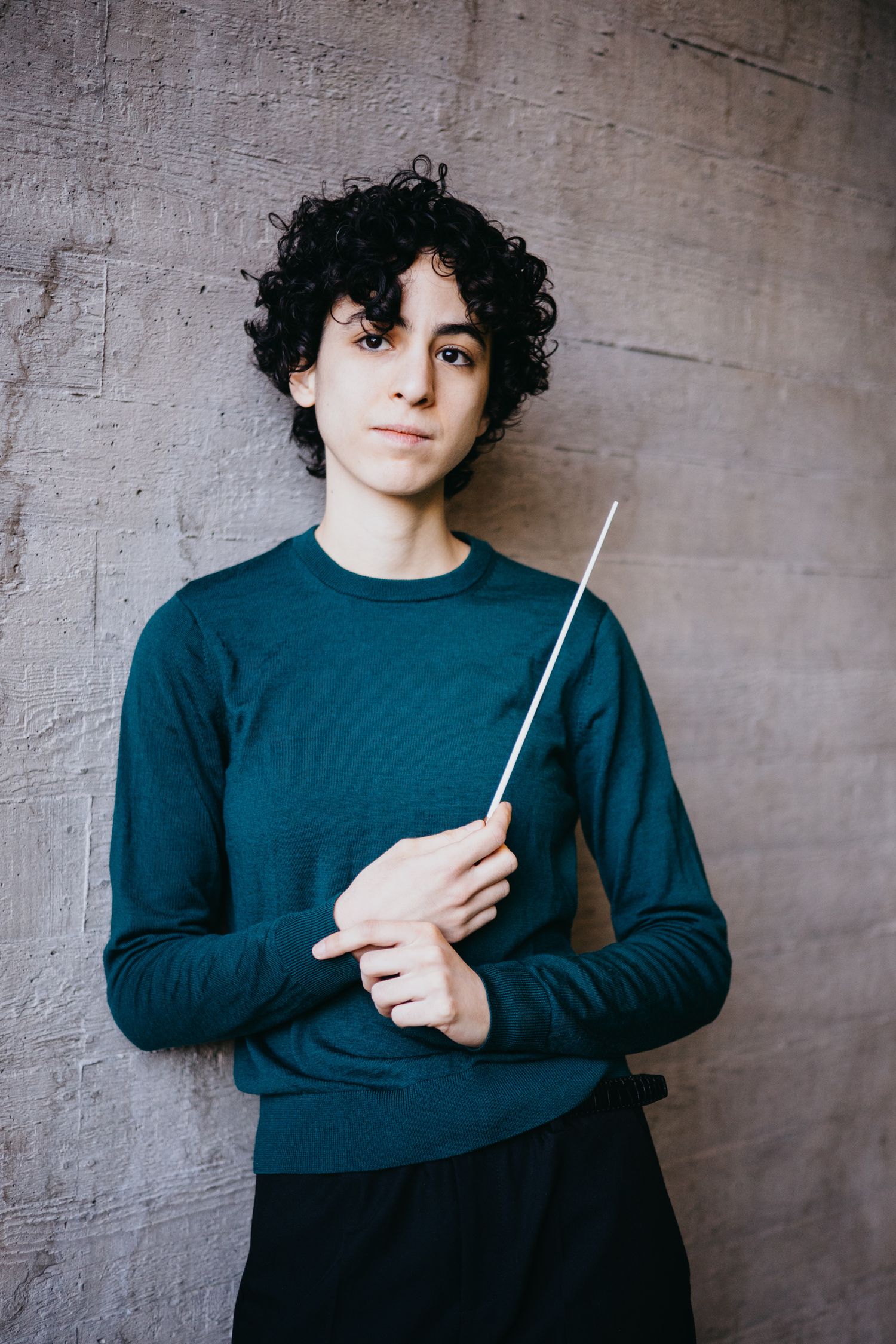 Young conductor holding a baton in teal sweater against concrete wall.