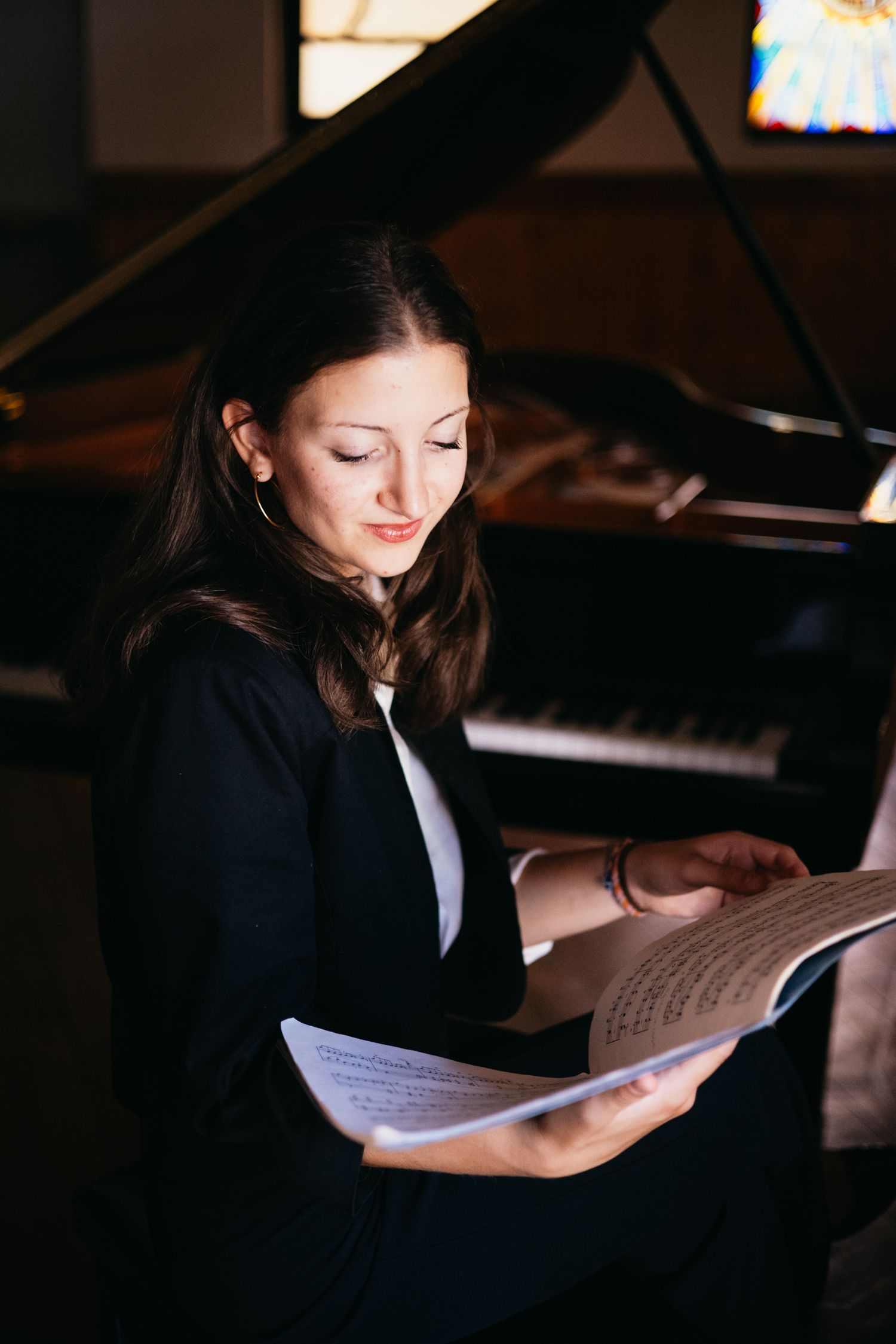 A young woman in a black blazer reads sheet music while seated in front of a grand piano in a dimly lit room.