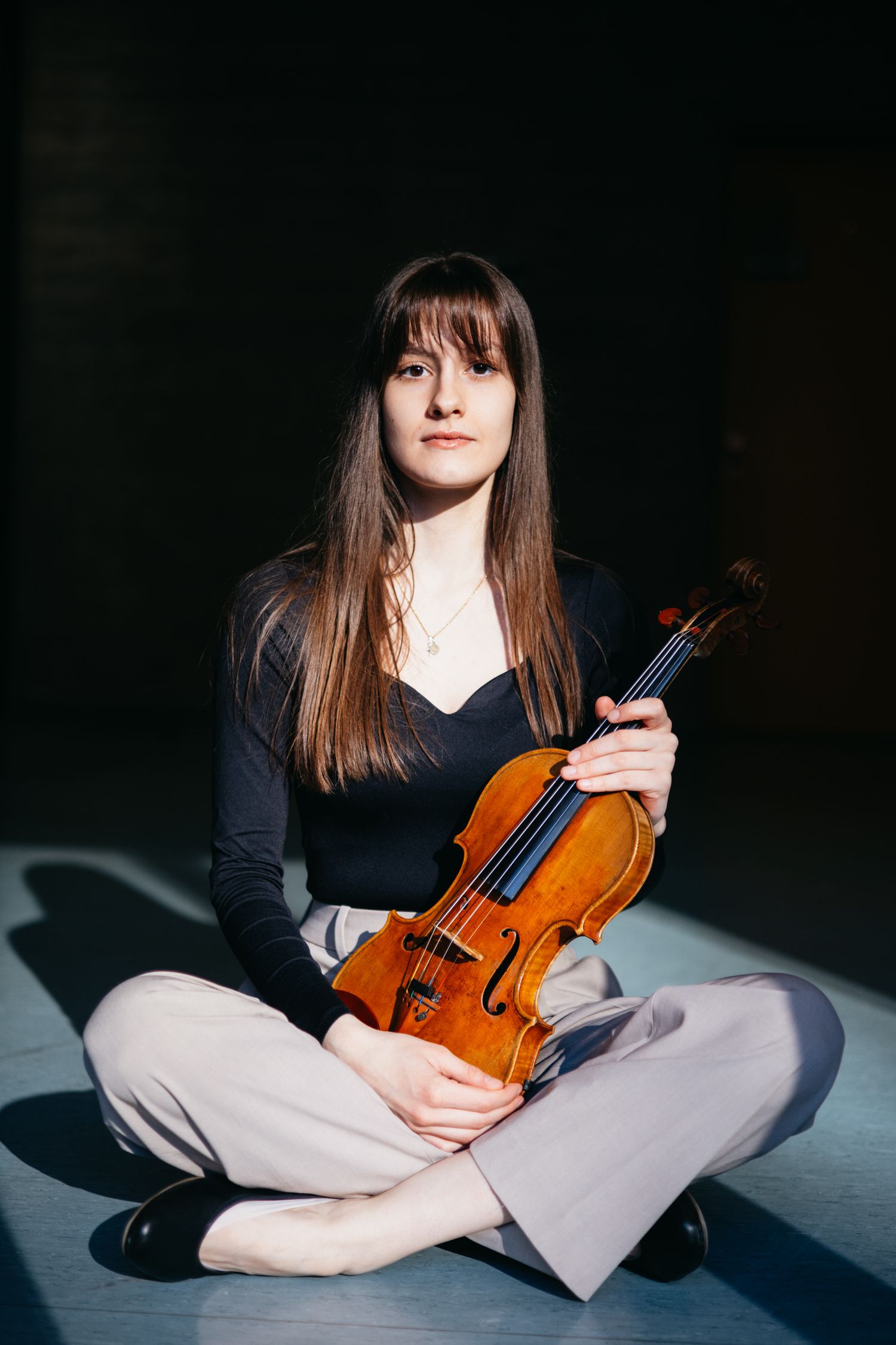 Young female violinist sits cross-legged on a studio floor holding her violin in dramatic light.