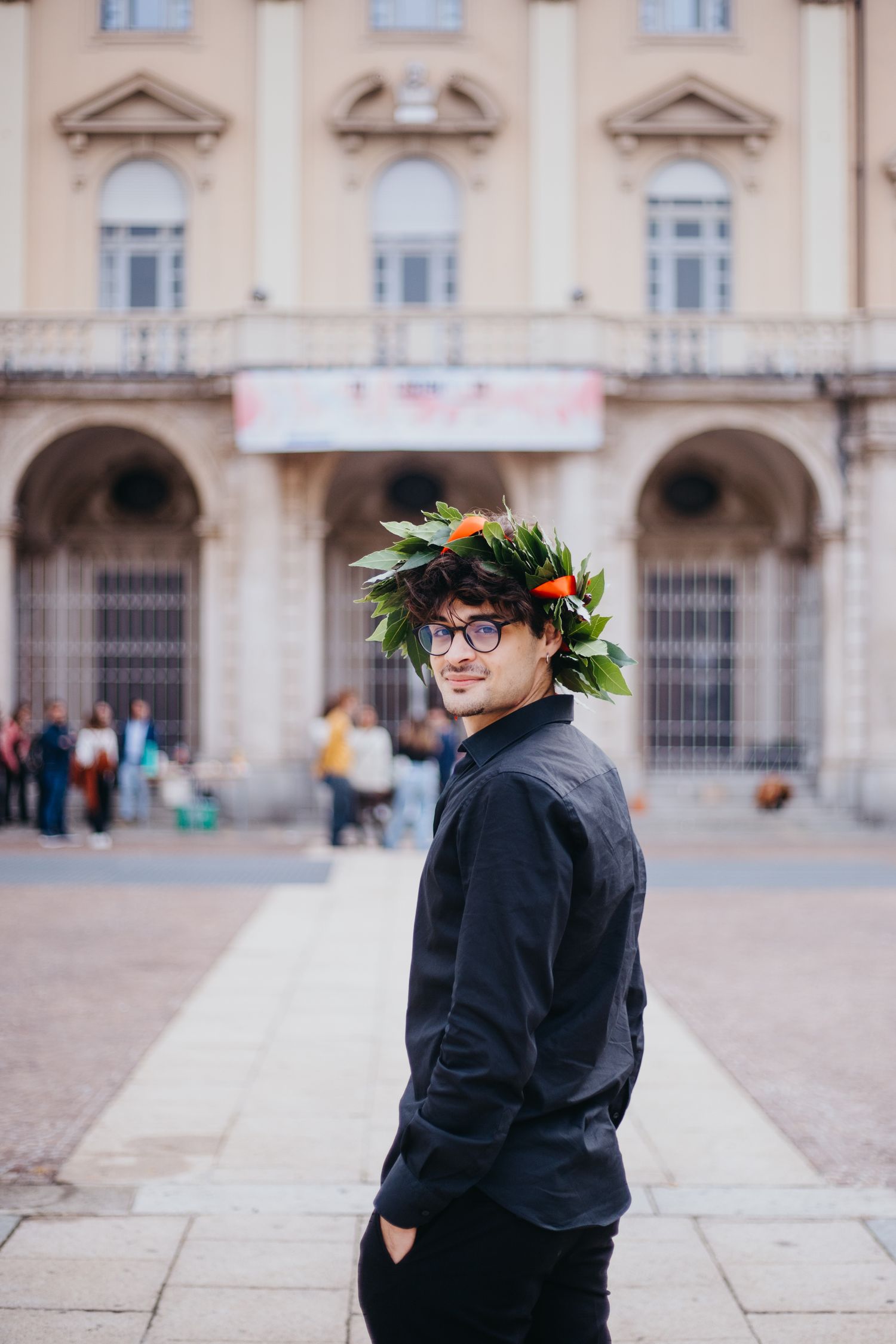 Young male graduate in laurel wreath poses outdoors in front of a grand classical building.