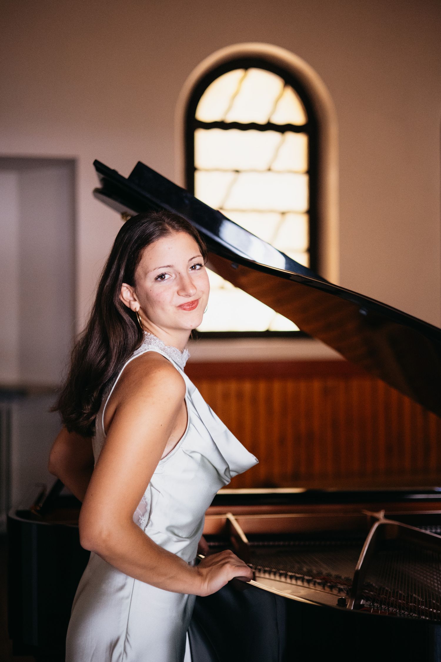 A young woman in a silver dress poses elegantly beside a grand piano in a concert hall with arched windows.