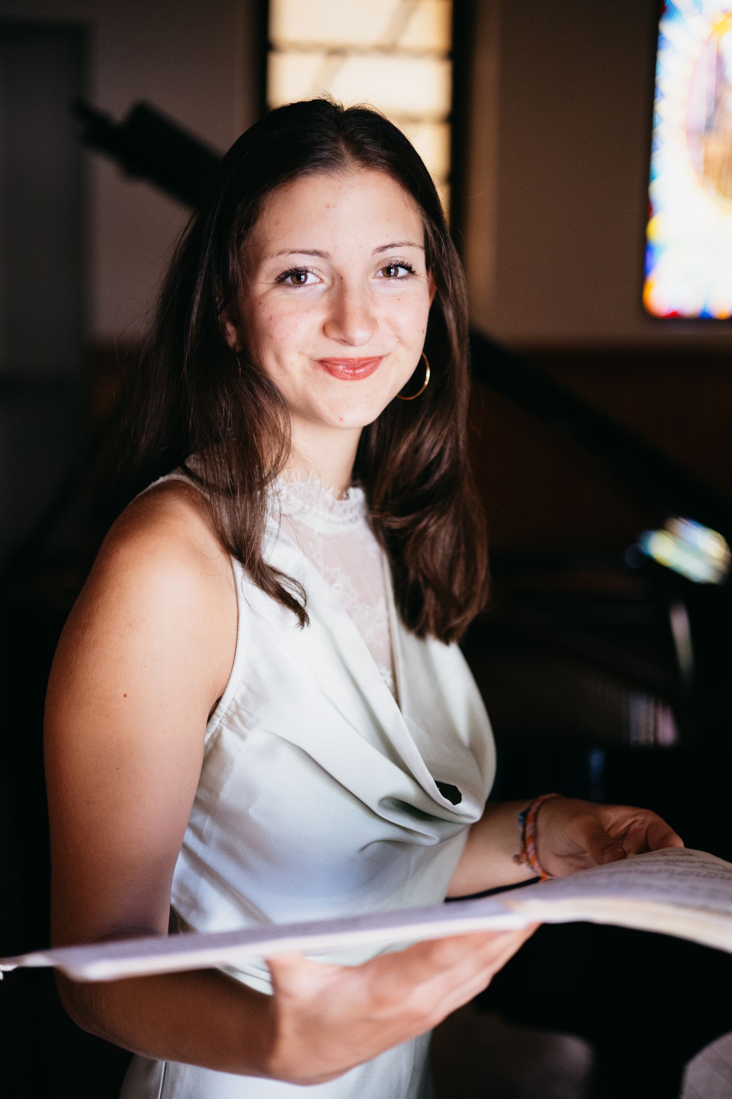Young woman in white dress holding sheet music, smiling in a church with stained glass window background.