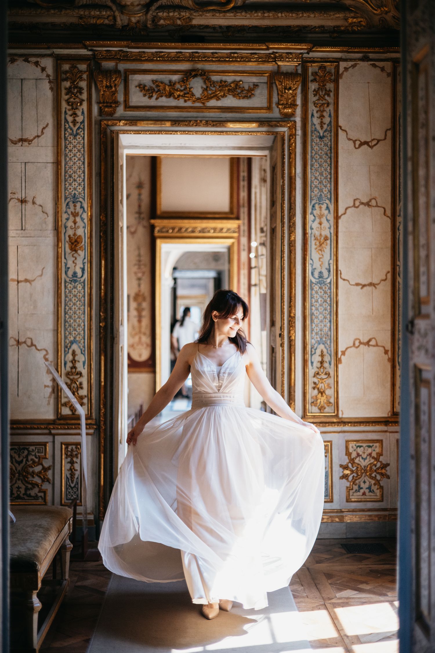 Bride in flowing white gown twirling in ornate golden baroque palace hallway with marble floors.