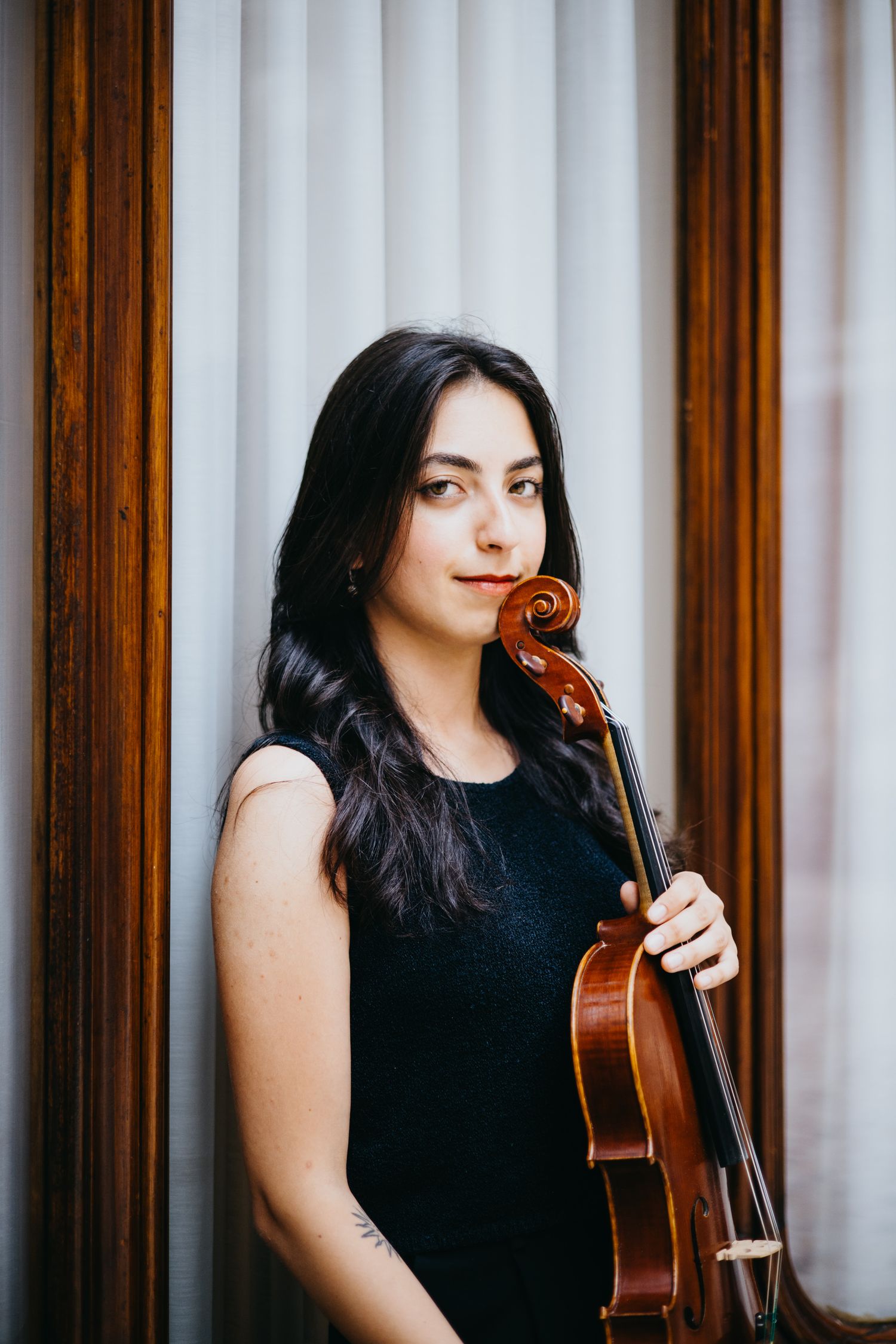 Young brunette female violinist in black dress holds violin near wooden framed window.