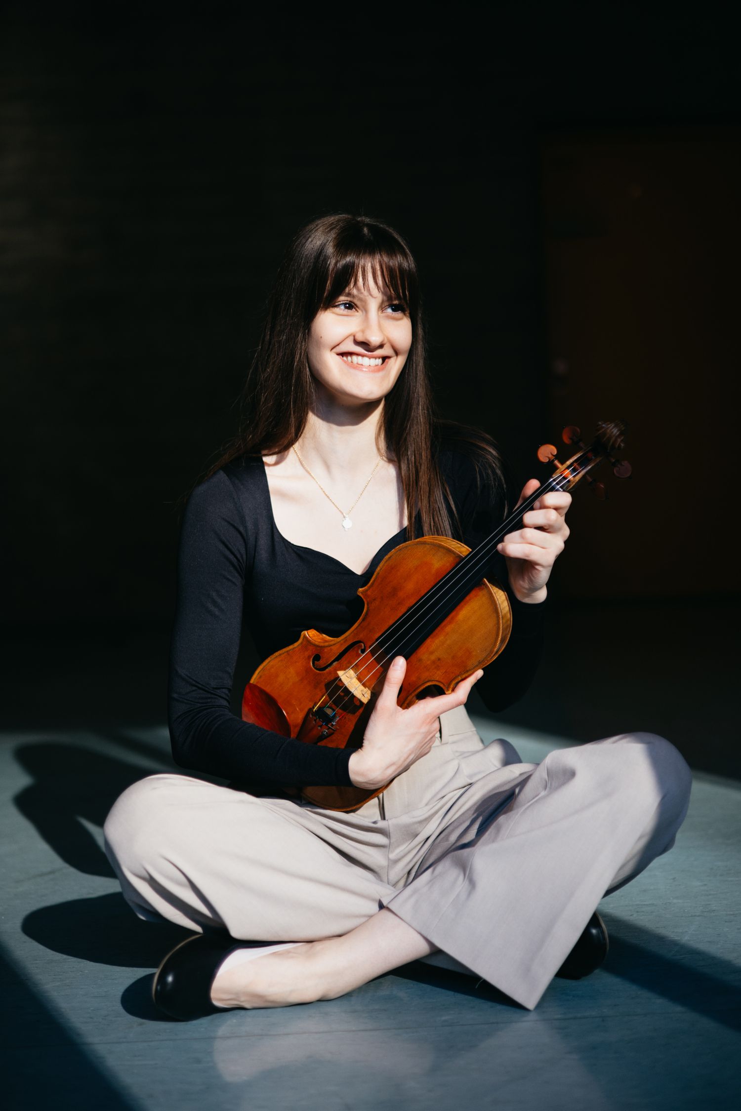 Smiling young female violinist sits cross-legged on floor holding her violin in warm spotlight.