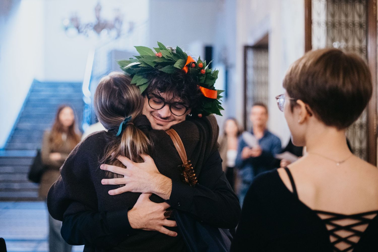 Smiling graduate wearing laurel wreath hugs guest at Italian university graduation ceremony indoors.